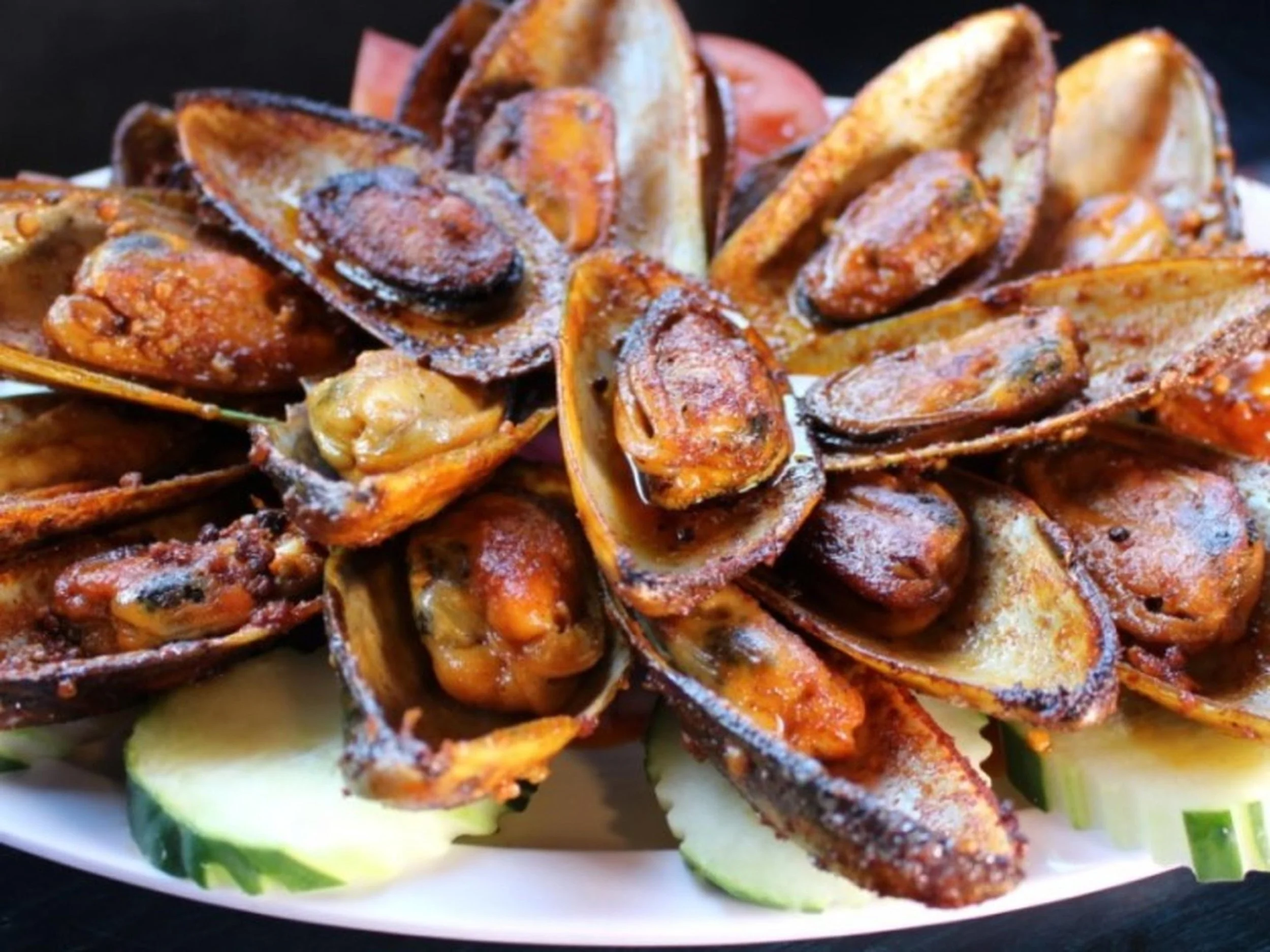 Grilled mussels served on a white plate with cucumber slices and a dark background.