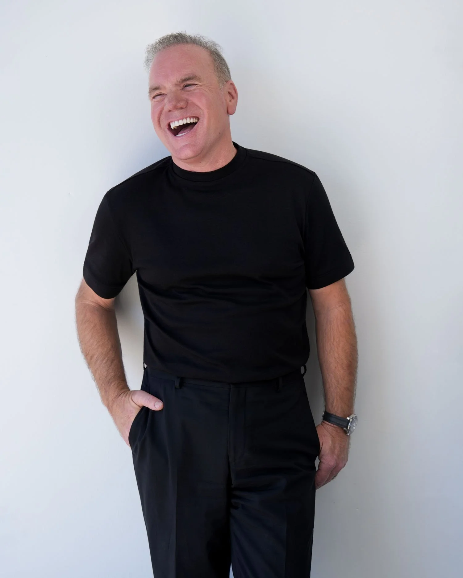 A smiling man with short light hair wearing a black t-shirt and black pants standing against a plain white wall.