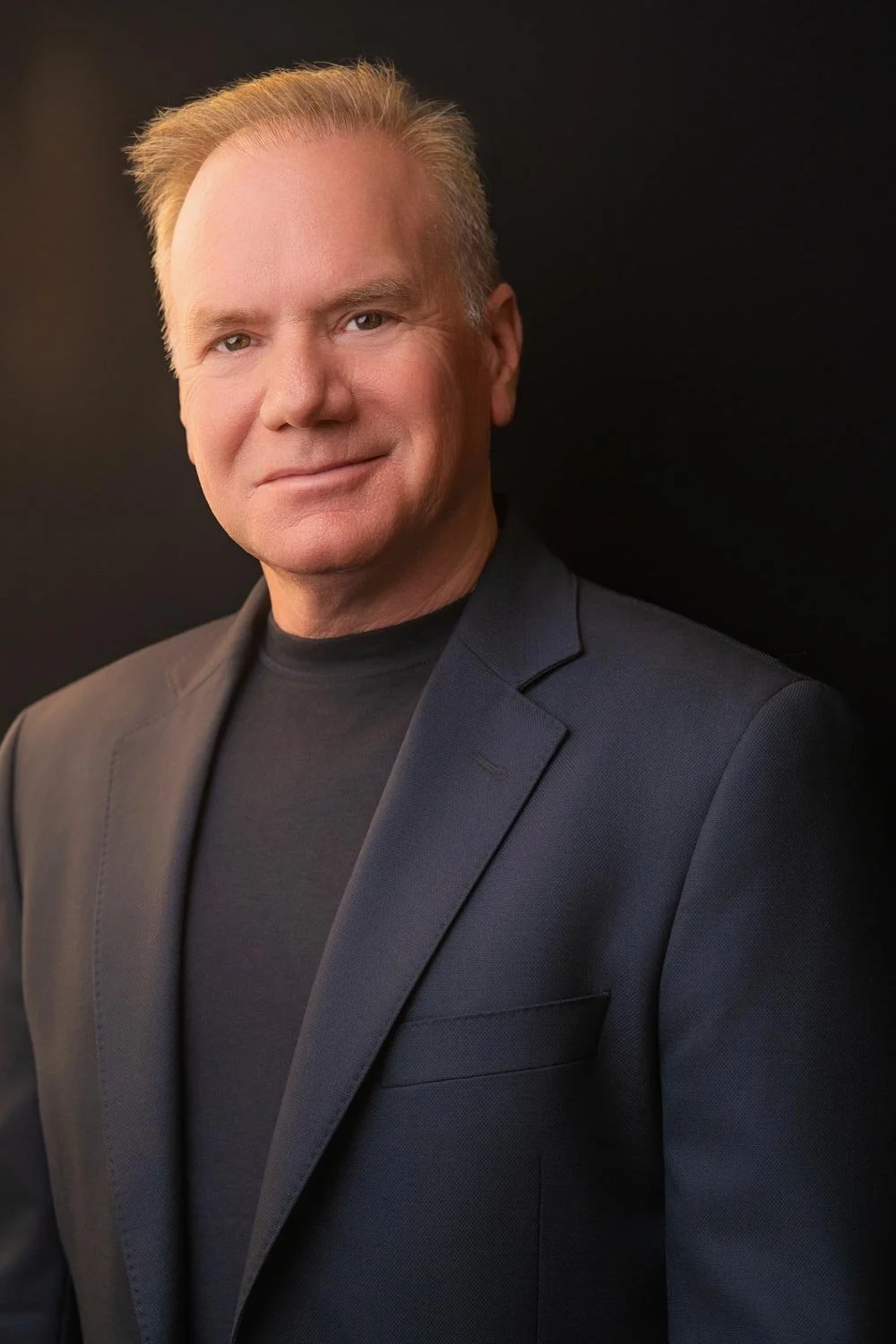 A close up portrait image of a man in a suit jacket. He is looking at the camera smiling. He has kind eyes.