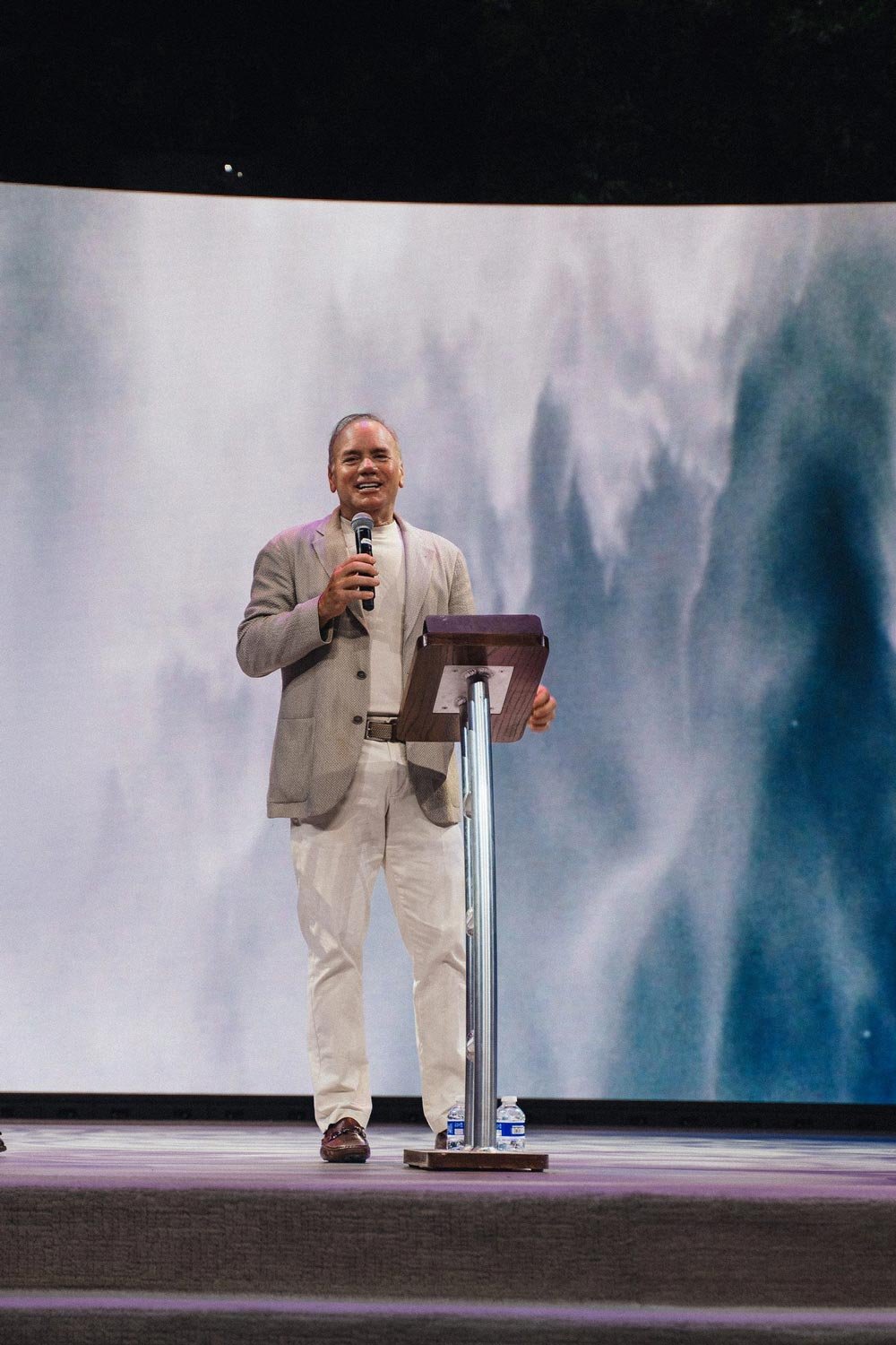 Rodney stands on stage in front of a podium with a microphone in his hand. He is speaking to a crowd with a smile on his face