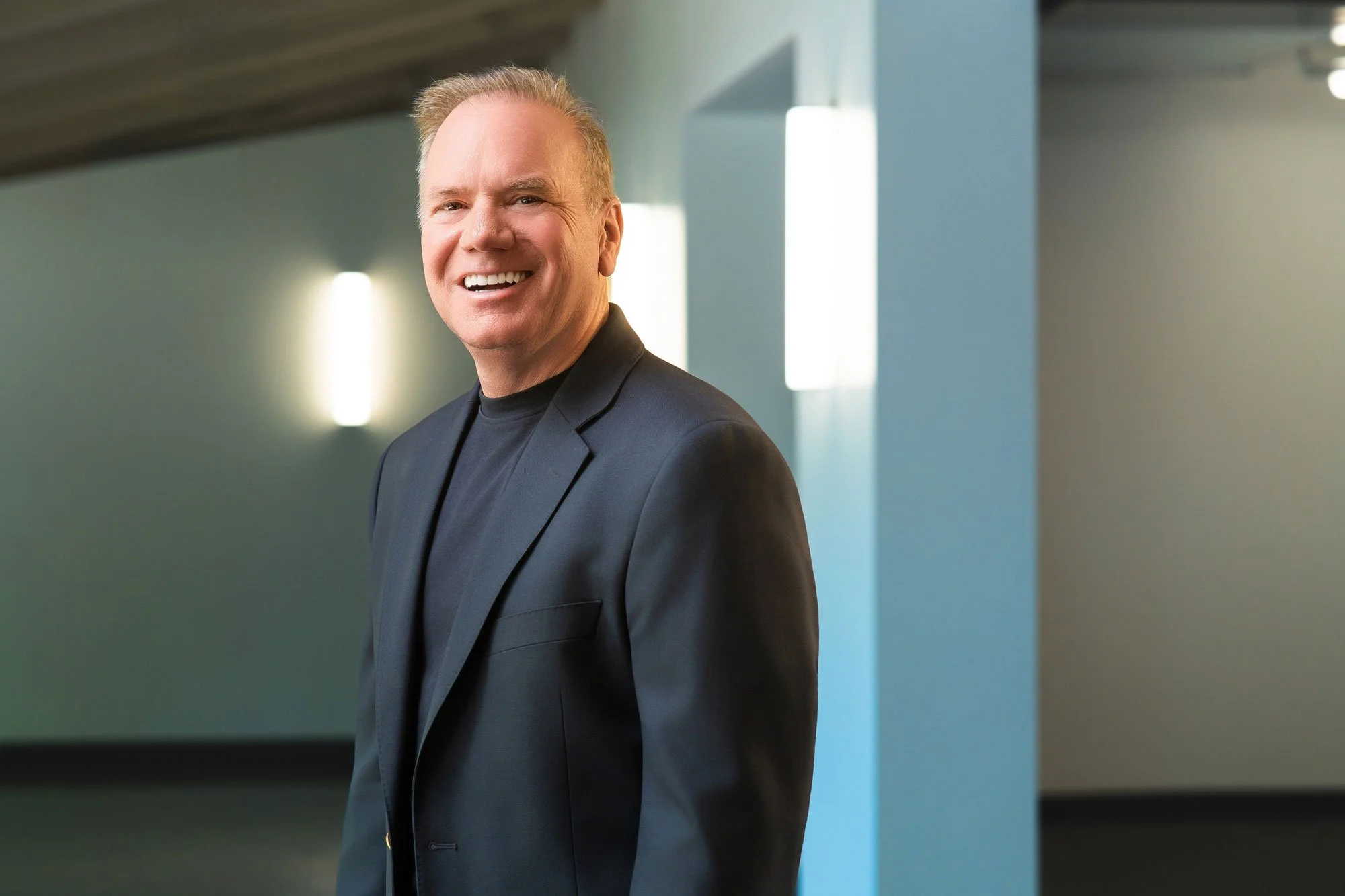 A smiling man with short light hair wearing a black blazer and black shirt standing in a modern indoor setting with soft lighting.