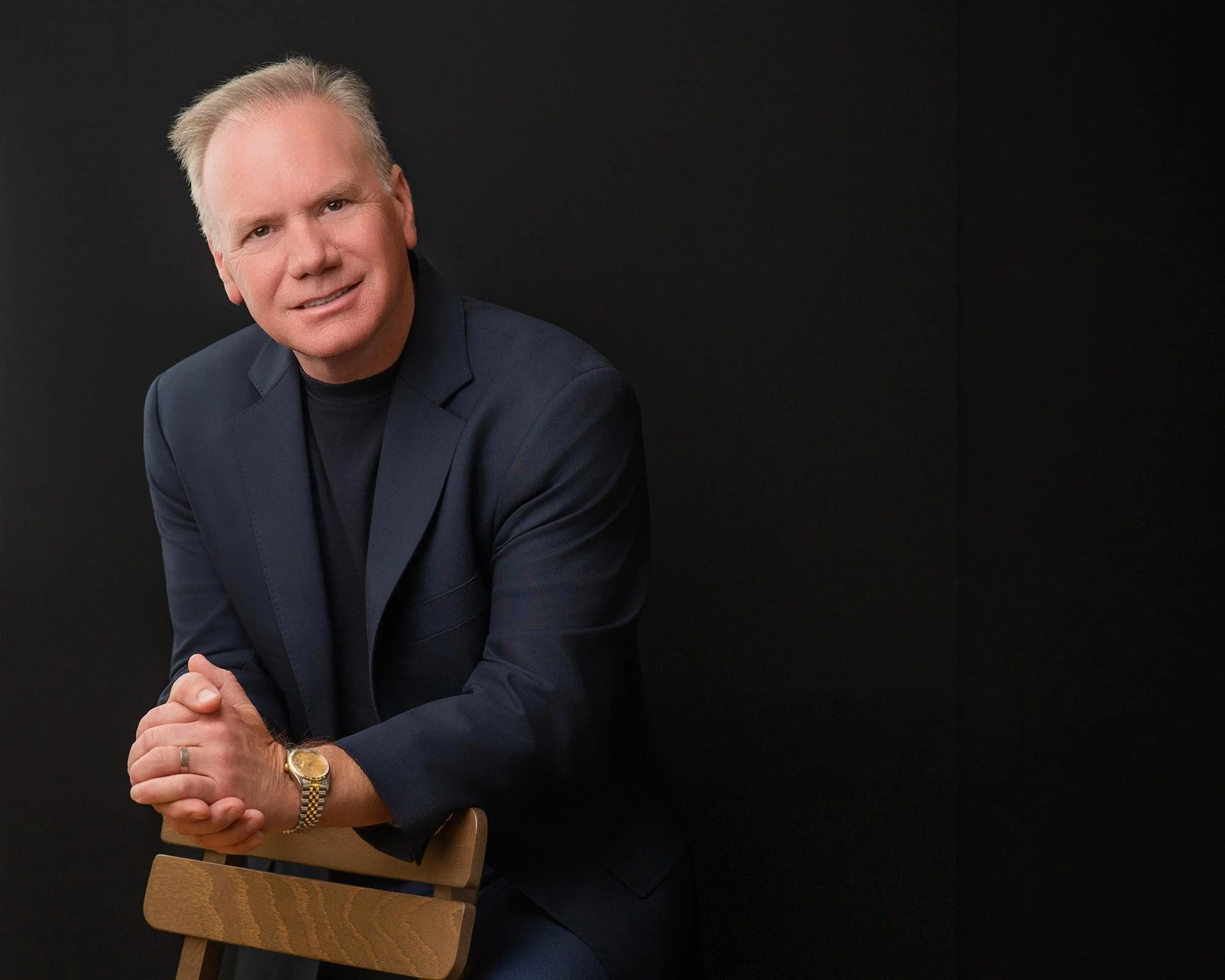 Rodney D. Nielsen, dressed in a dark blue blazer and black shirt, has his arms over a wooden chair against a black background. He is smiling slightly, with his hands clasped together, wearing a gold watch and a gold ring on his left hand.