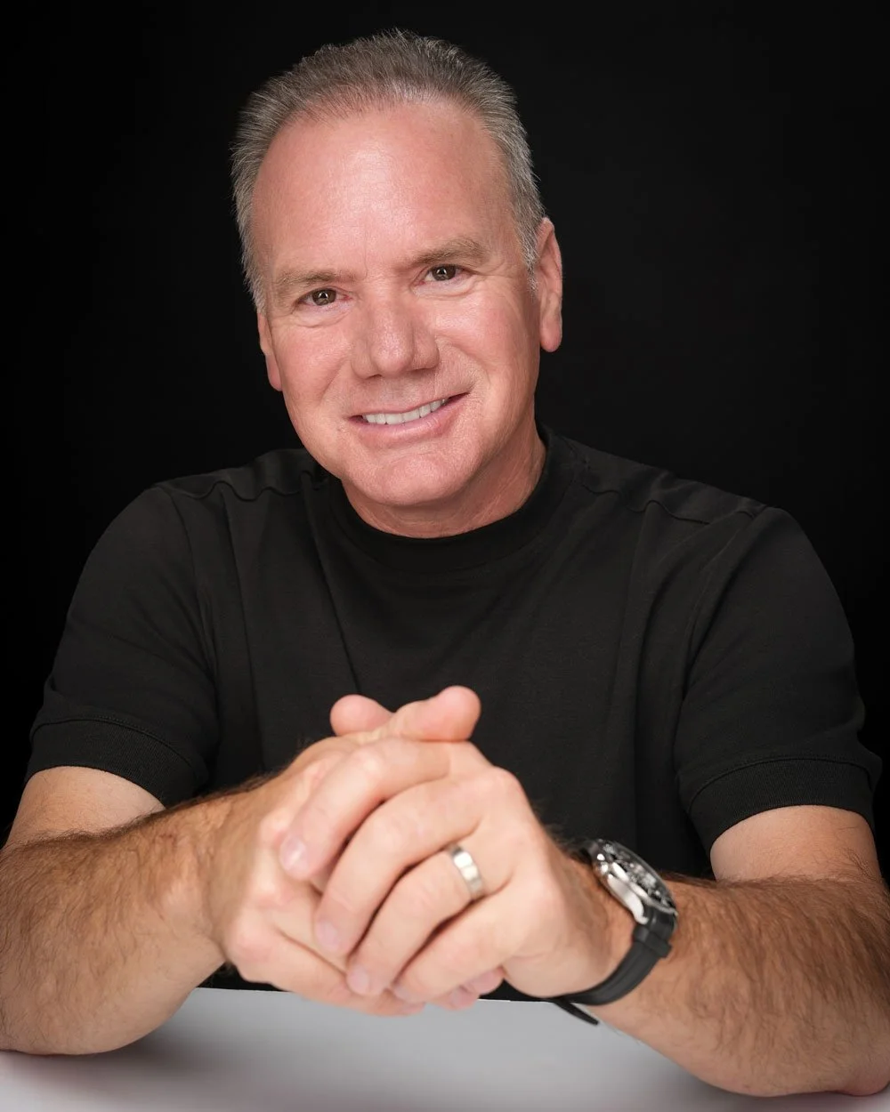 Rodney D. Nielsen smiles. He is a man with short grey hair, wearing a black T-shirt, sitting with his hands clasped on a white table, against a black background.