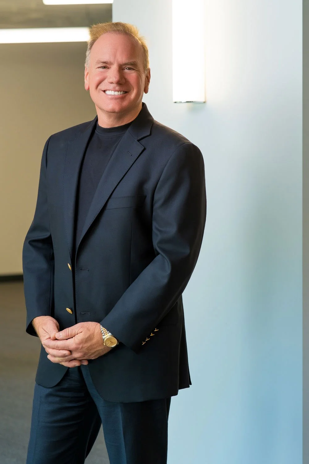 A smiling man in a black suit standing indoors with a modern light fixture on the wall behind him.