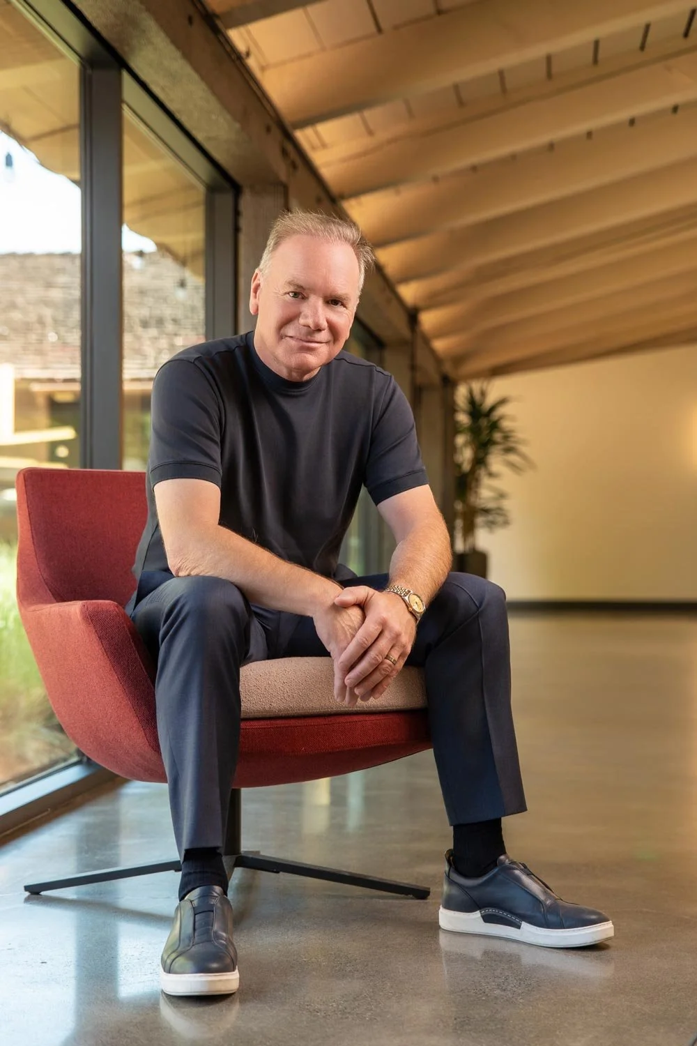A man sitting on a red and beige chair indoors near large glass windows with a wooden ceiling overhead. He is wearing a black T-shirt, dark pants, black sneakers, and a wristwatch, smiling at the camera.