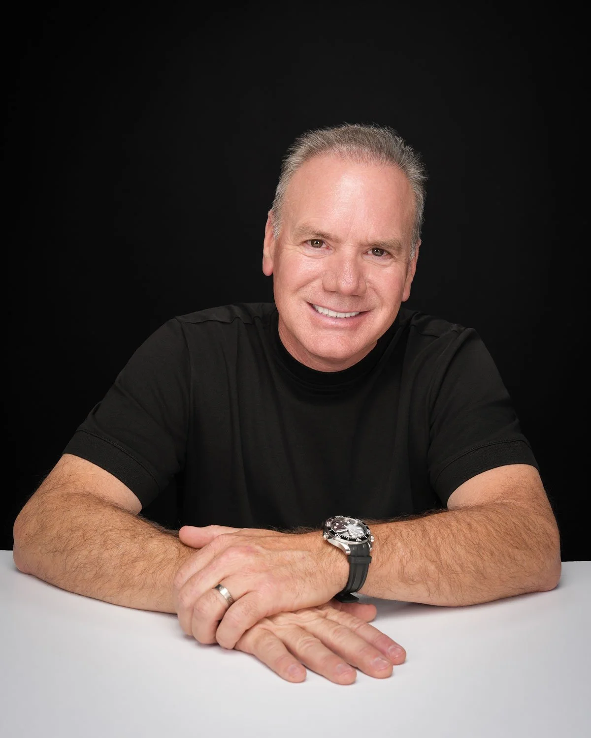 Rodney sits at a white table, he sits with his hands on the table smiling and looking at the camera