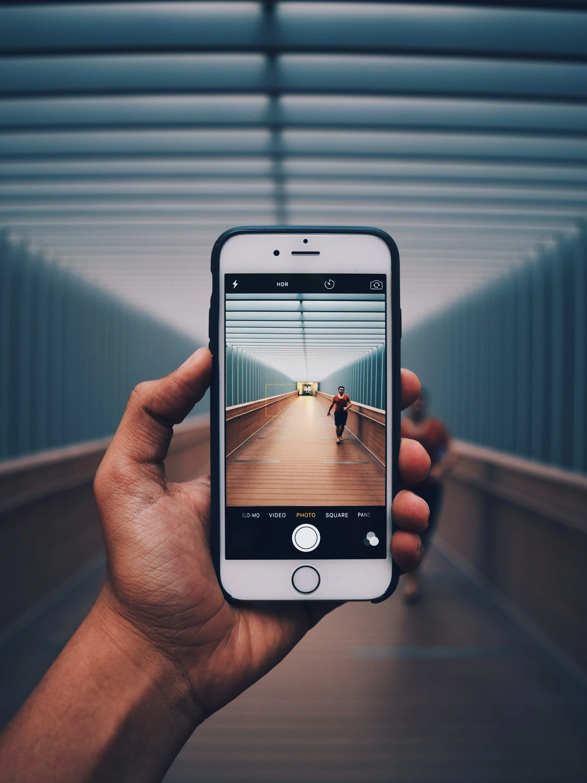 Hand holding a smartphone taking a picture of a person walking in an underground tunnel with blue walls and a wooden floor.