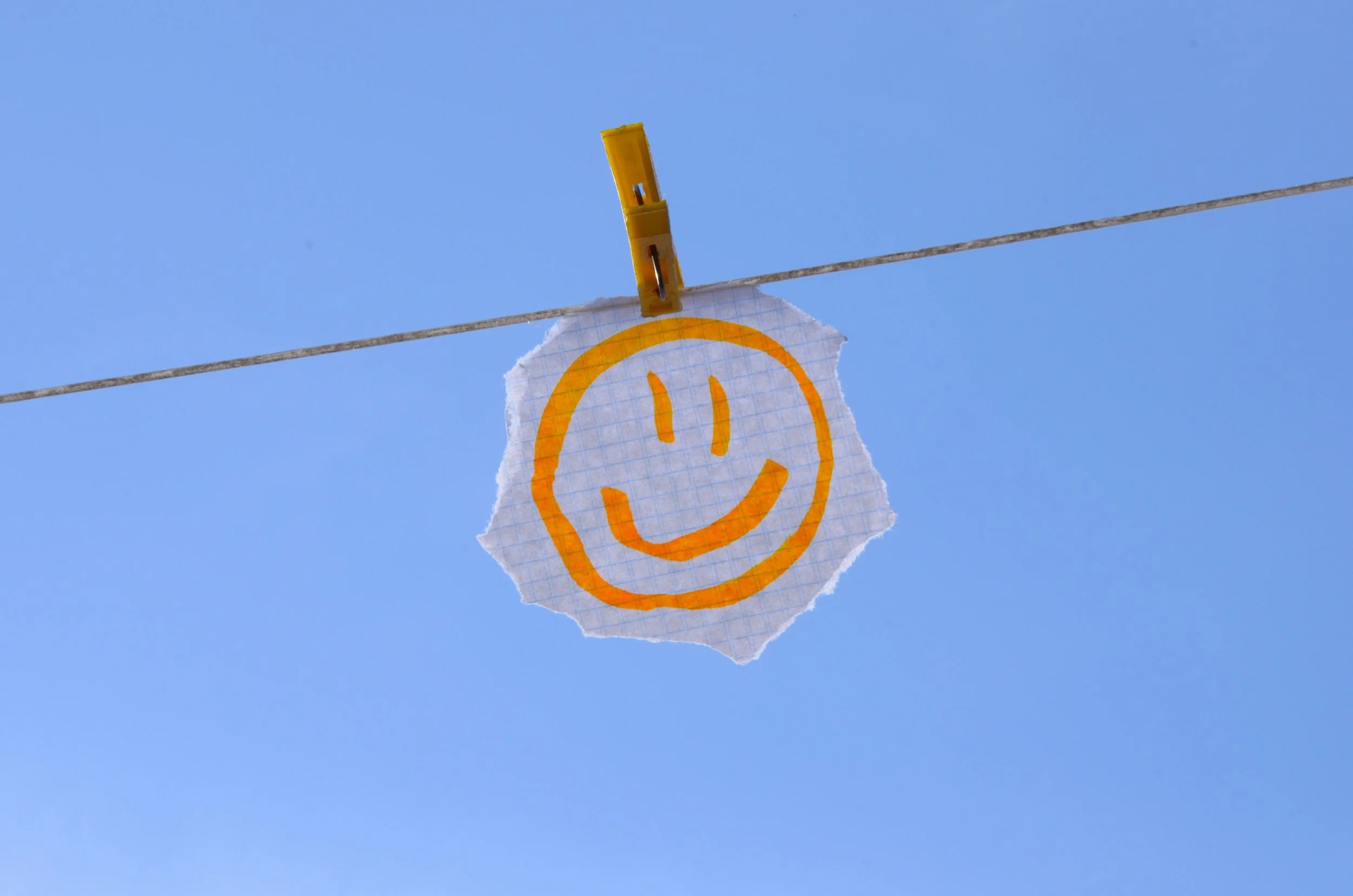 A piece of torn graph paper with a yellow smiley face drawn on it, clipped onto a clothesline with a yellow clothespin, against a clear blue sky.