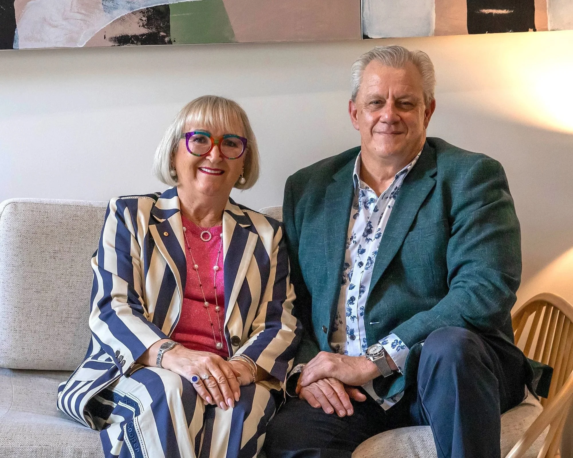 A woman with blonde hair wearing colourful glasses, a navy and white striped blazer, and jewelry, smiling; seated next to a man with gray hair, also smiling, dressed in a patterned shirt and teal blazer, holding hands, in a cozy indoor setting.