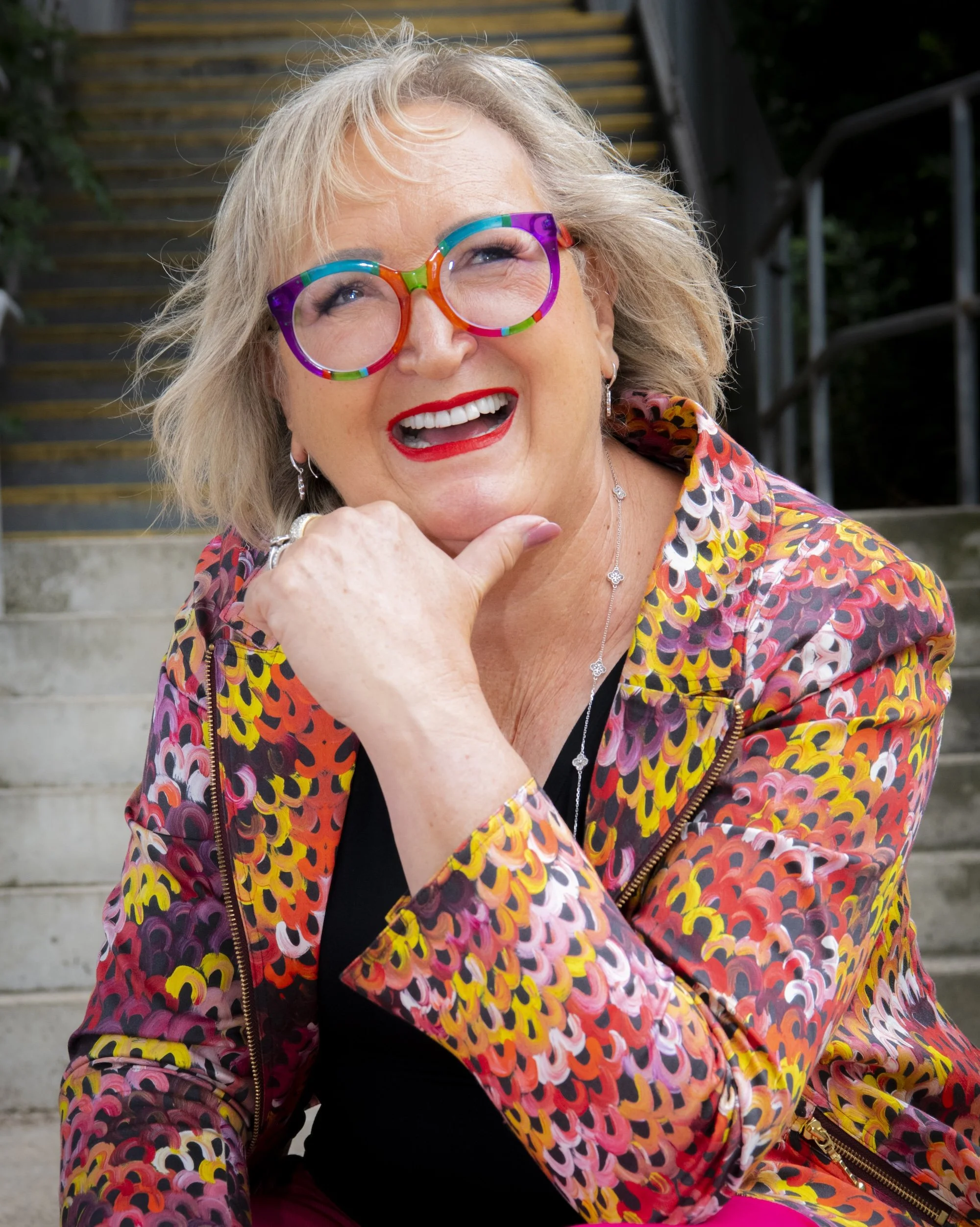 A woman with blonde hair wearing colourful, patterned glasses and a vibrant jacket, smiling and sitting on outdoor stairs.