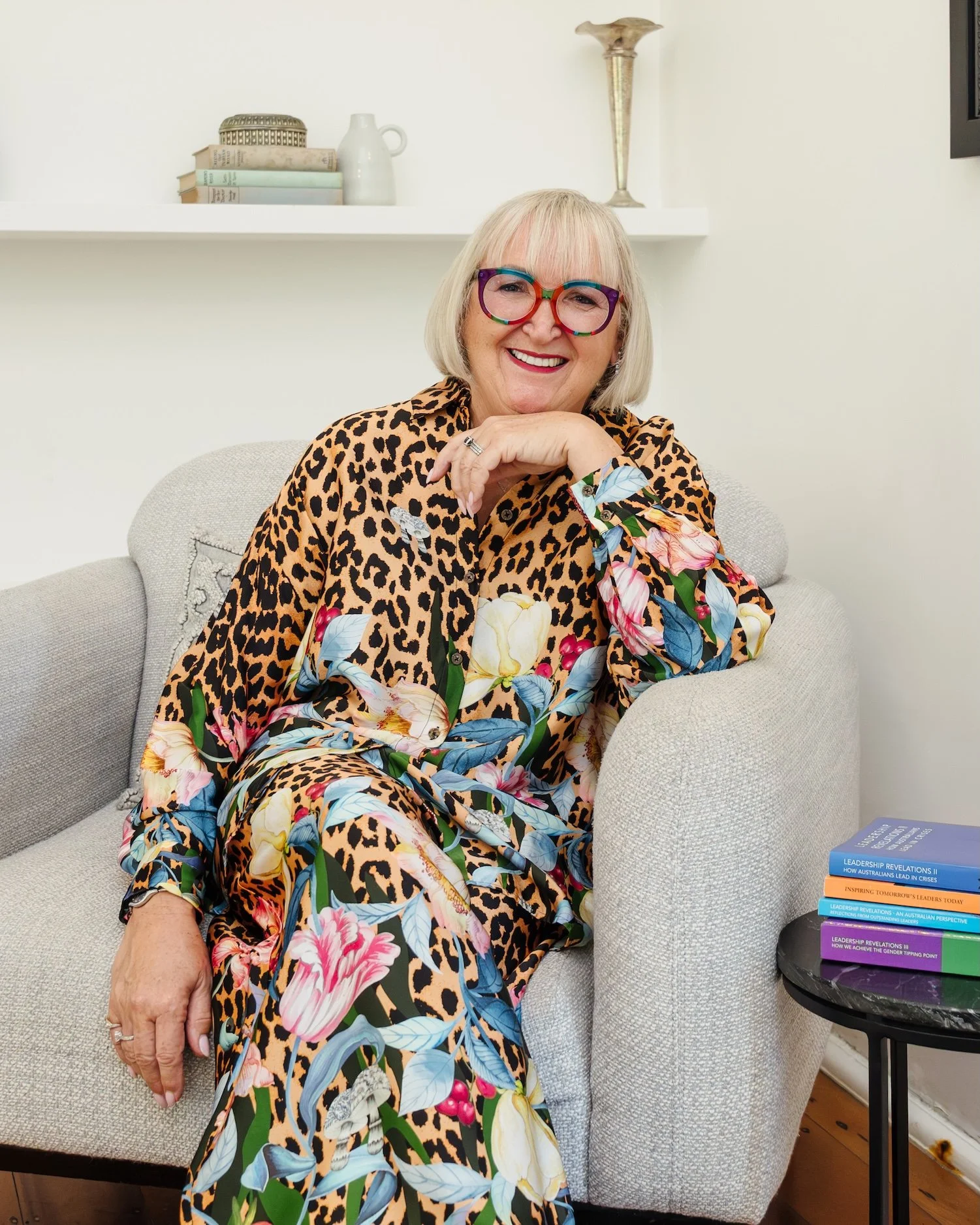 A smiling woman with blonde hair and colorful glasses, wearing a leopard print and floral dress, seated on a light-colored couch in a cozy room with a bookshelf and a small table with books.