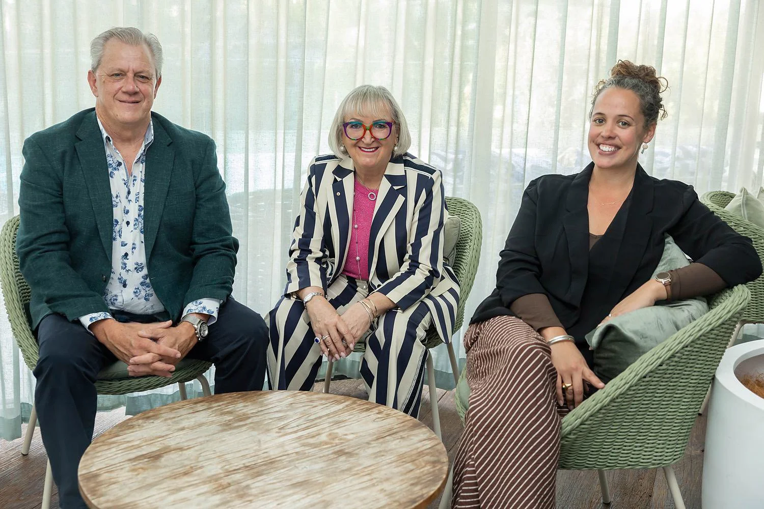 Three people sitting on chairs with a round table in front of them; a man in a dark blazer and floral shirt, a woman in a striped suit with glasses and jewelry, and a woman in a black blazer and striped pants, with a light-colored curtain in the background.
