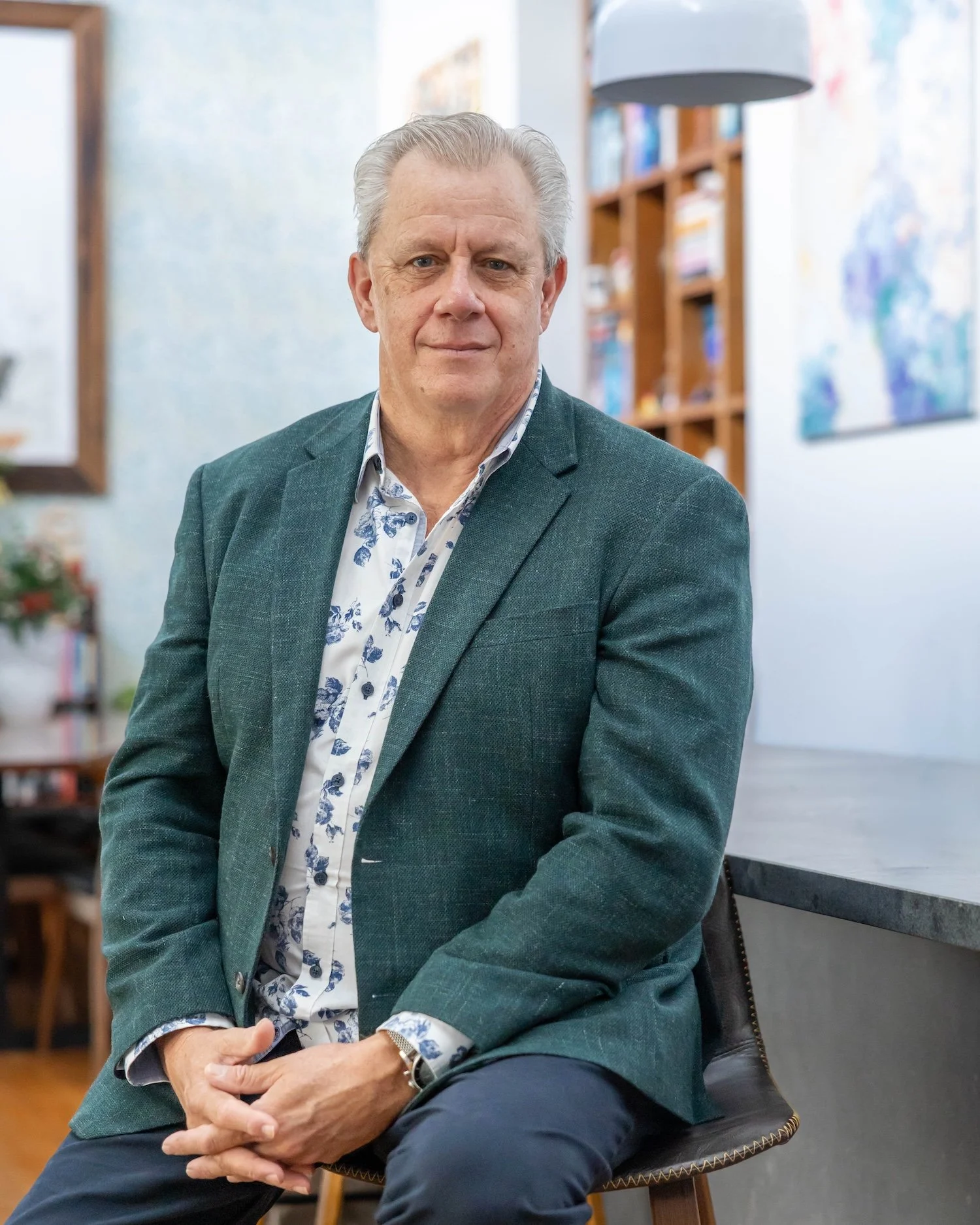 An serious man sitting on a chair in an indoor setting, wearing a dark green blazer over a white shirt with a blue pattern, with a bookshelf and colourful artwork in the background.