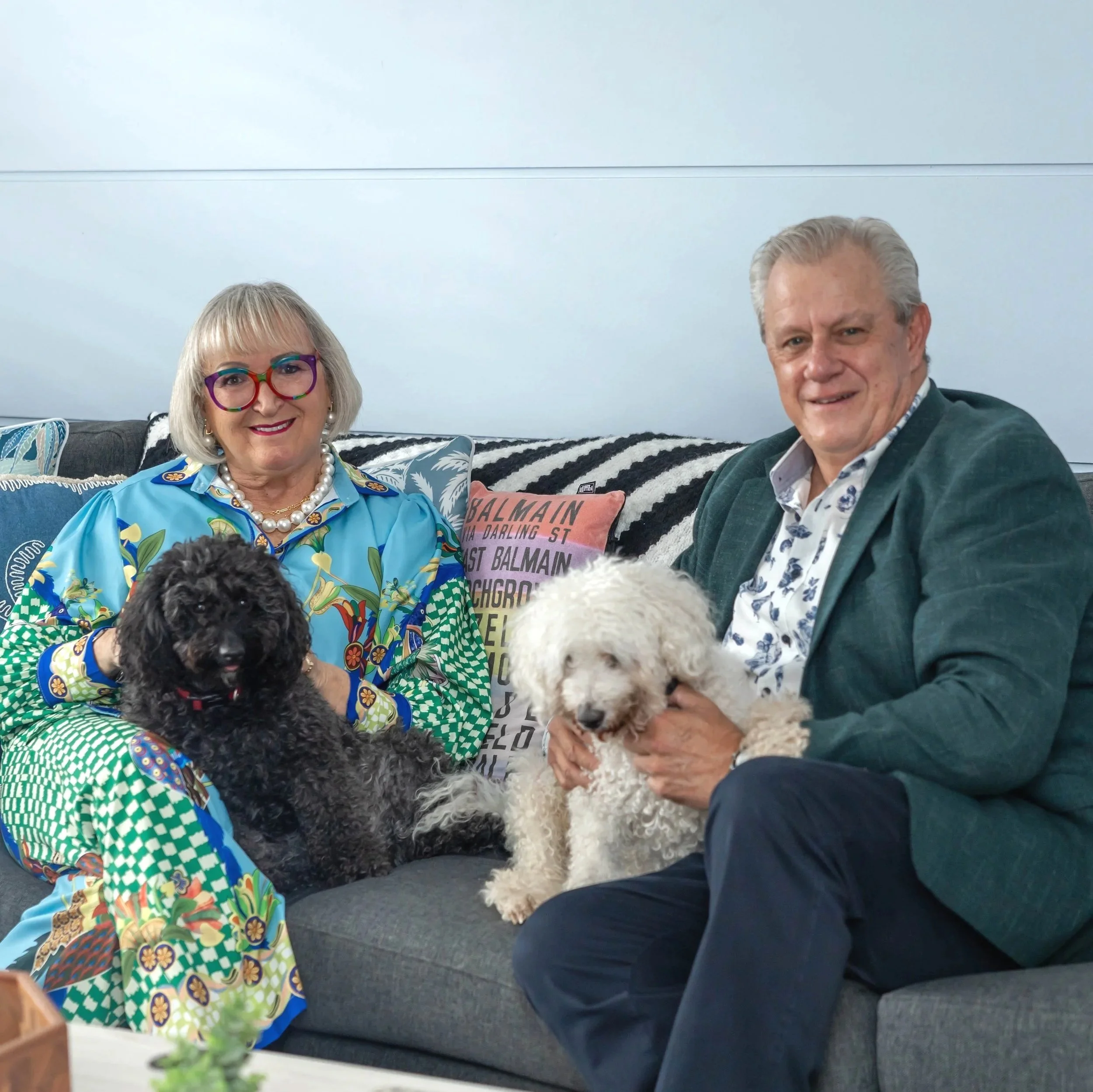 A woman with blonde hair, wearing colorful glasses and a bright blue shirt with floral and geometric patterns, sitting on a sofa with a black and white striped pillow. Next to her is a man with gray hair, in a dark blazer and white shirt, sitting on the sofa. They are holding fluffy poodles, one black and one white. The background features a minimalist light gray wall and a cushion with the word 'Balmain' on it, with a plant visible in the foreground.