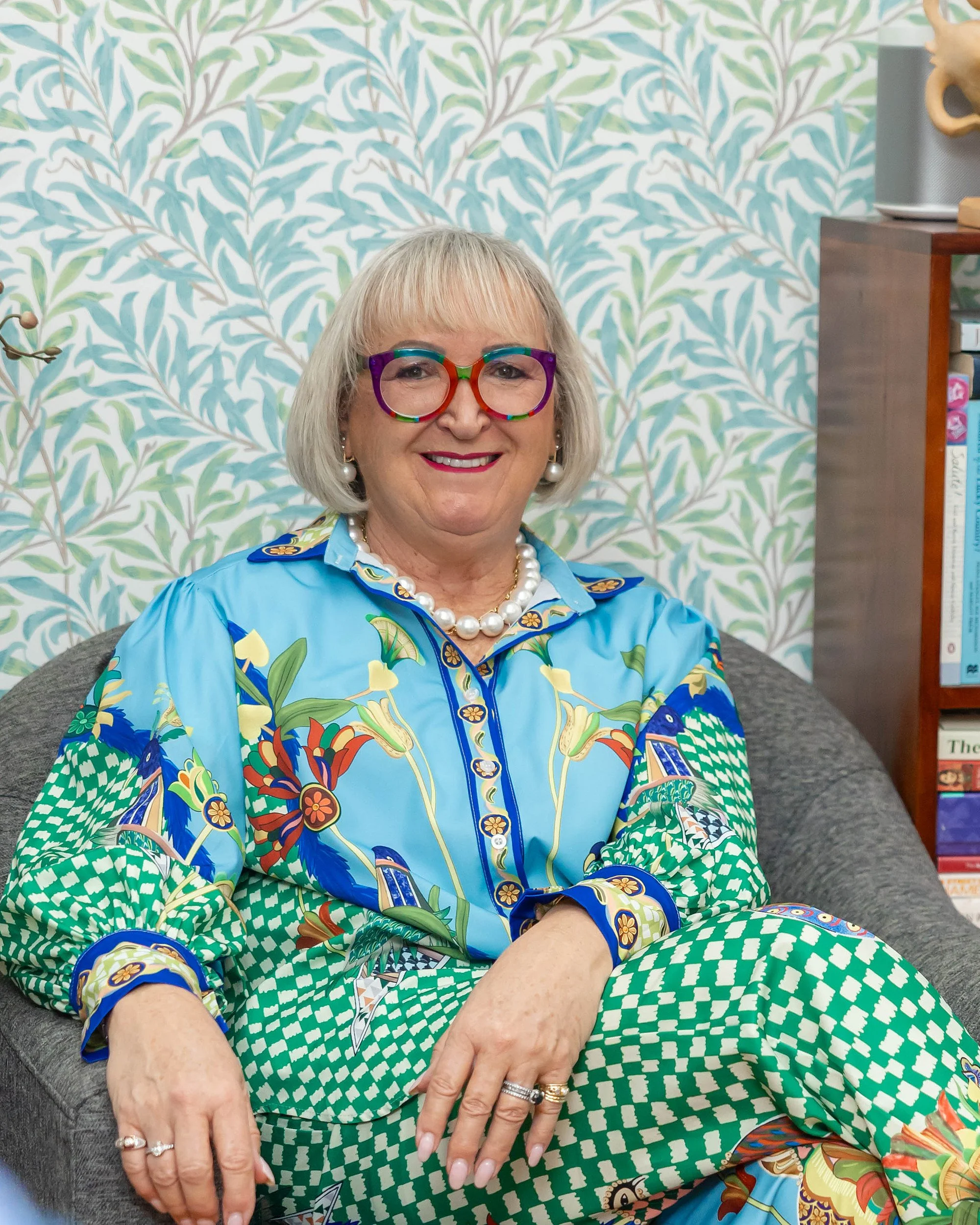 A smiling woman with blonde hair and colourful glasses, wearing a vibrant, patterned shirt and matching pants, sitting on a gray couch against a leafy patterned wallpaper background, with bookshelves partially visible in the background.