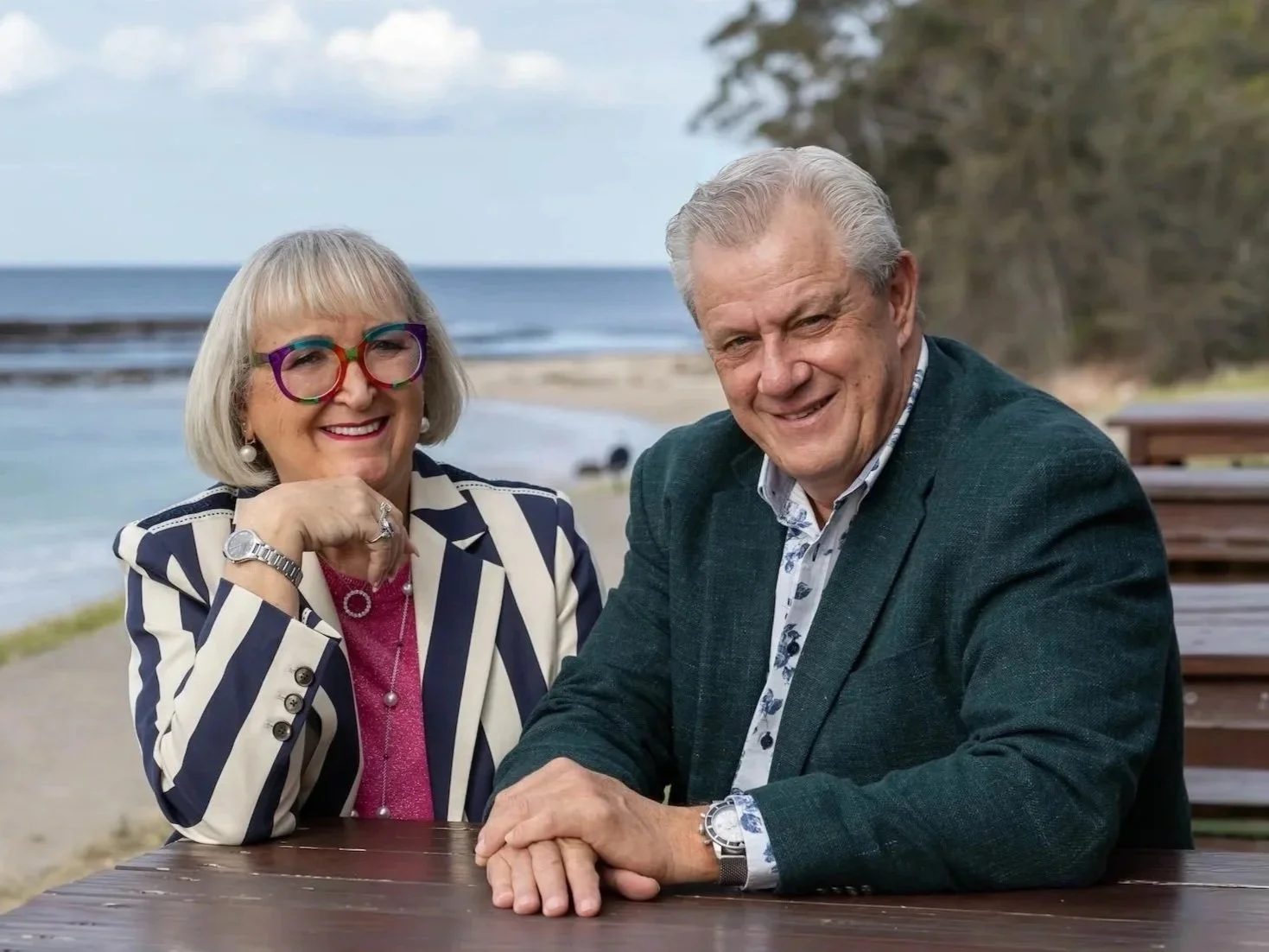 A smiling couple sitting at a table outdoors near the beach. The woman is wearing colourful glasses, a striped blazer, and jewelry. The man is wearing a dark blazer and a patterned shirt. The background shows the ocean.