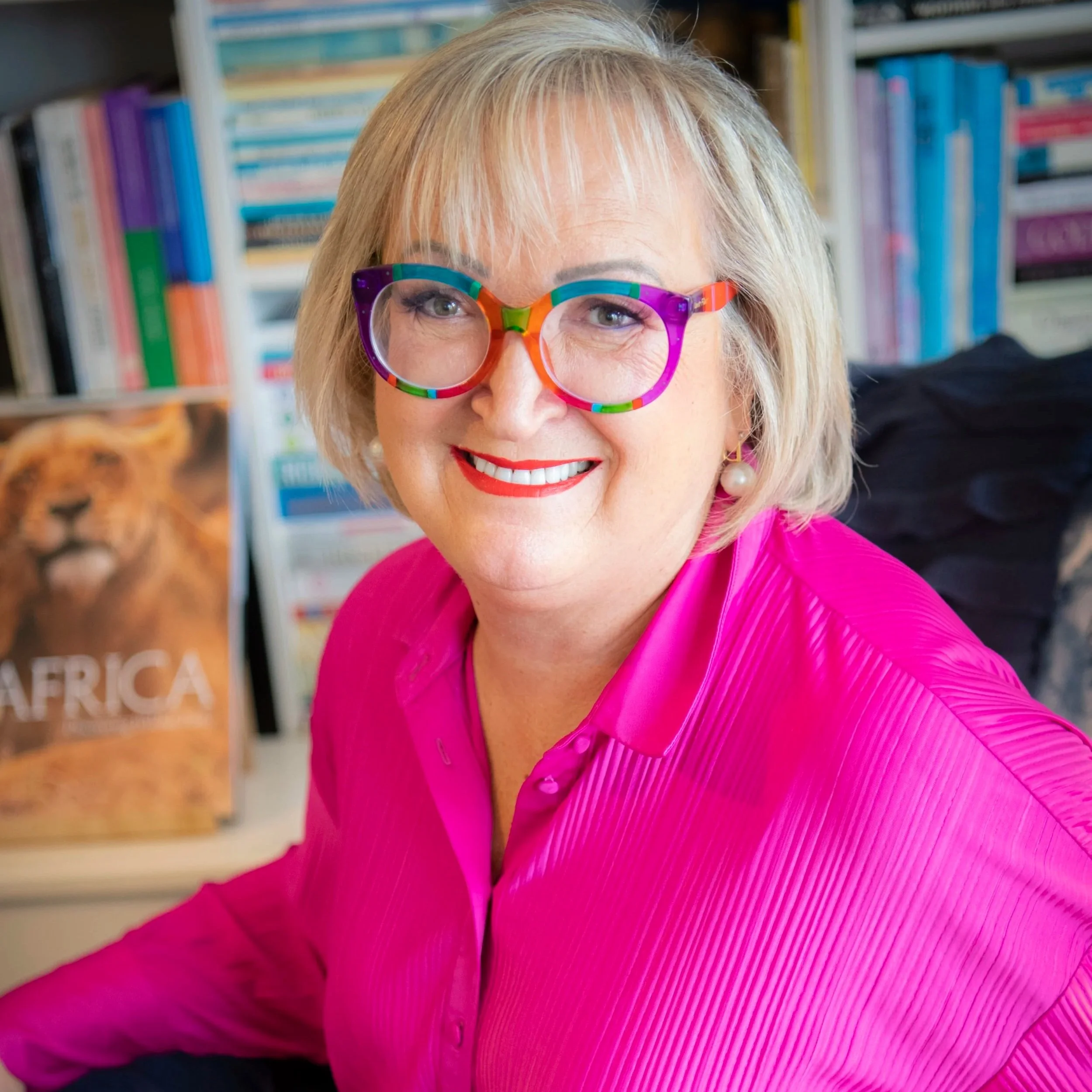 Close-up of a woman with blonde hair wearing colourful glasses and a bright pink blazer, smiling in front of a bookshelf.