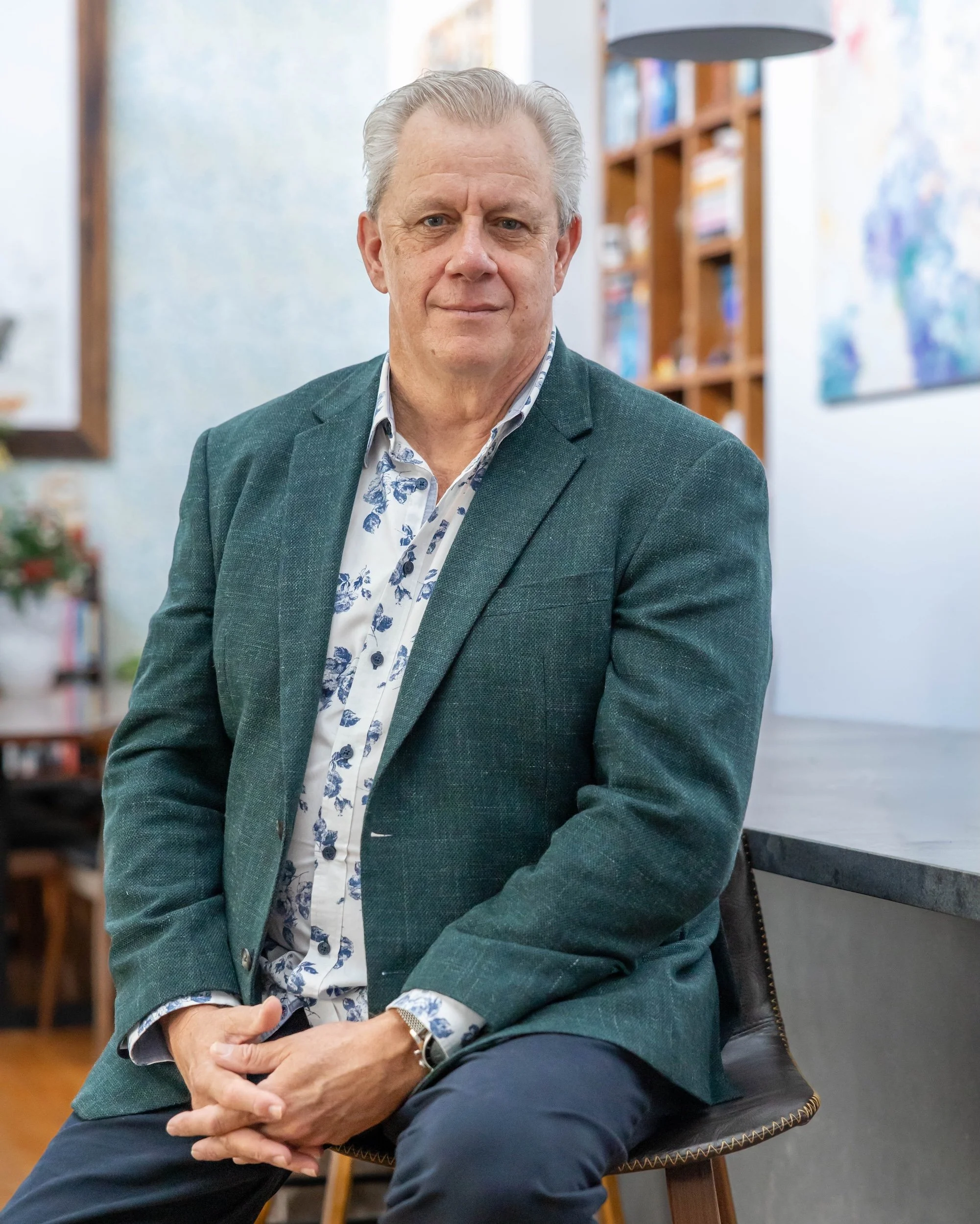 A serious man with gray hair, wearing a dark green blazer and a blue-and-white patterned shirt, sitting on a chair in a cozy, well-lit indoor space with bookshelves in the background.