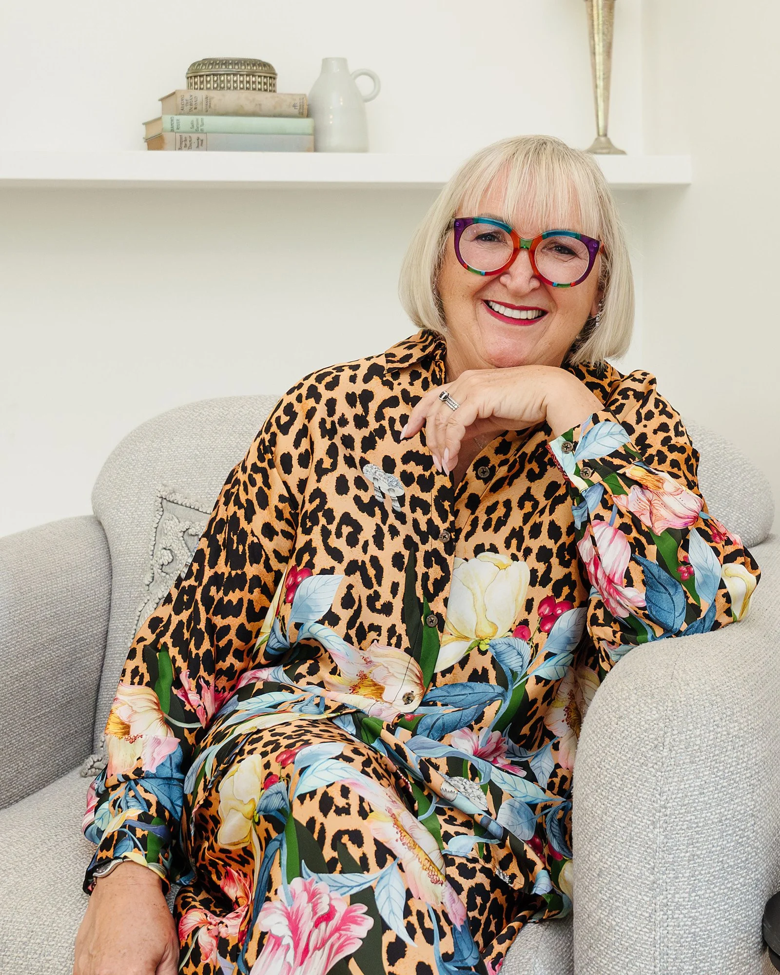 A smiling woman with blonde hair, wearing colorful leopard print and floral glasses, sitting on a light-coloured sofa with shelves of books, a vase, and decorative objects in the background.