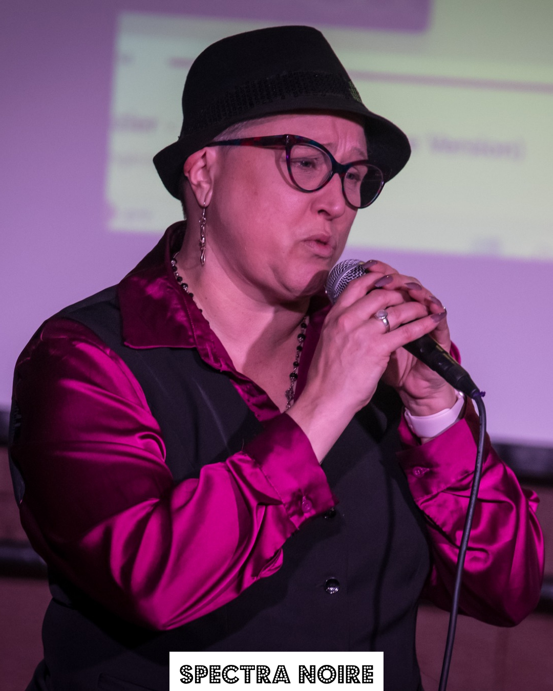 Woman wearing a black hat, glasses, and a shiny pink and black jacket, holding a microphone and speaking.