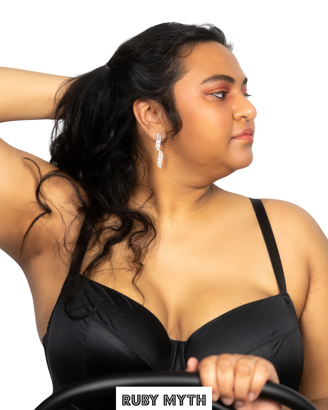A woman with dark wavy hair wearing glittery earrings and a black top, posing with her left hand behind her head and her right hand on a steering wheel, against a white background. The text 'RUBY MYTH' is at the bottom of the image.