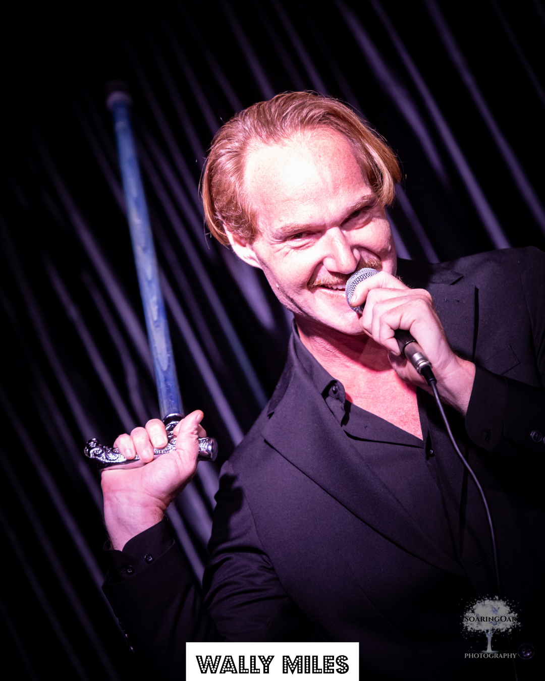 A man with red hair and a beard, wearing a black suit, holding a microphone in his right hand and a sword in his left, smiling on stage with a dark background and vertical light streaks.