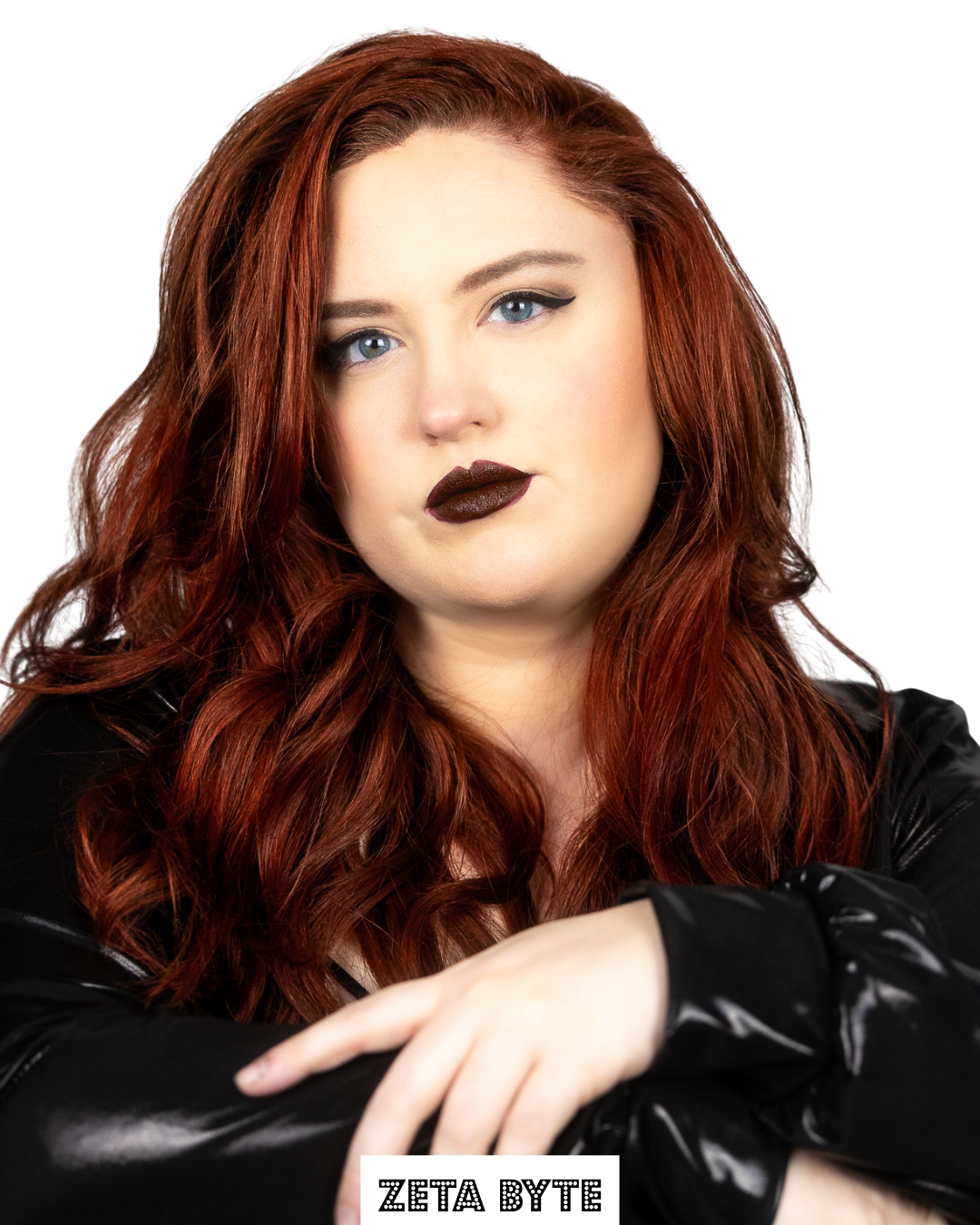 A woman with long, wavy red hair and blue eyes, wearing dark makeup and a black leather jacket, looking directly at the camera with her arm crossed in front of her and a serious expression.