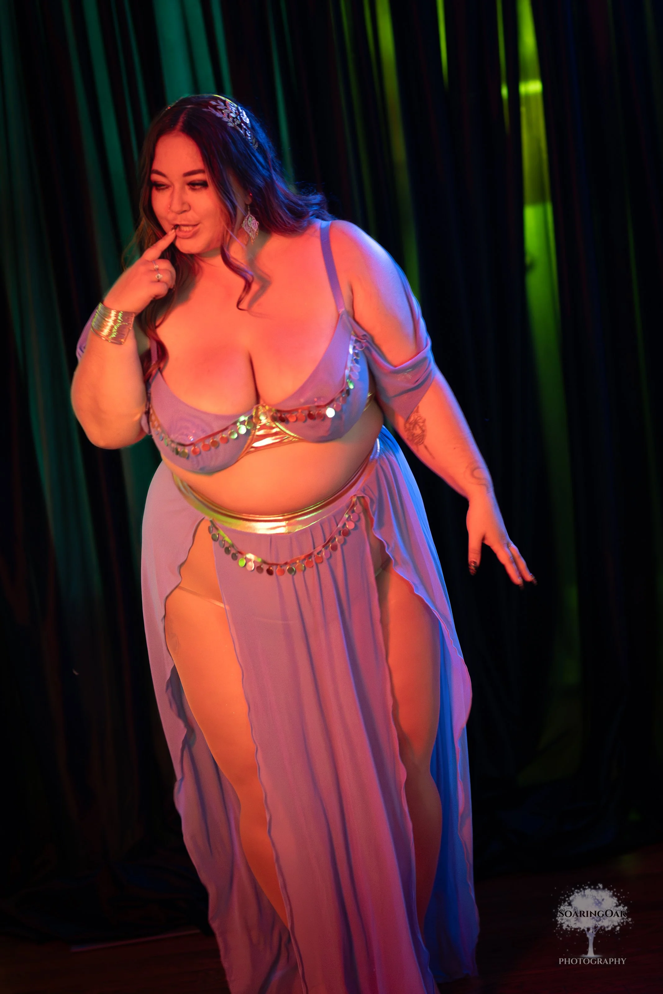 A woman with dark brown wavy hair and tattoos, wearing a lavender and colorful sequined dress, is dancing on stage with dark curtains in the background. She is touching her lips with one finger, looking down with a playful expression, and has jewelry including earrings and a bracelet.