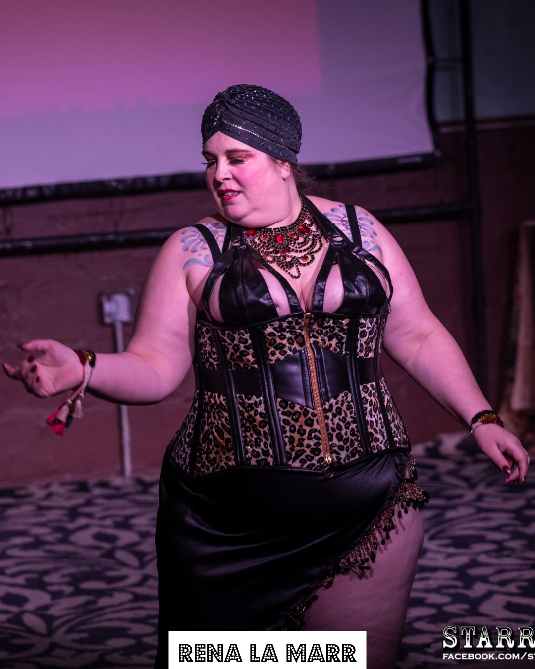 A woman wearing a black hat with rhinestones, a leopard-print and black leather corset, and a black skirt with fringe, performing on stage with a pink background.