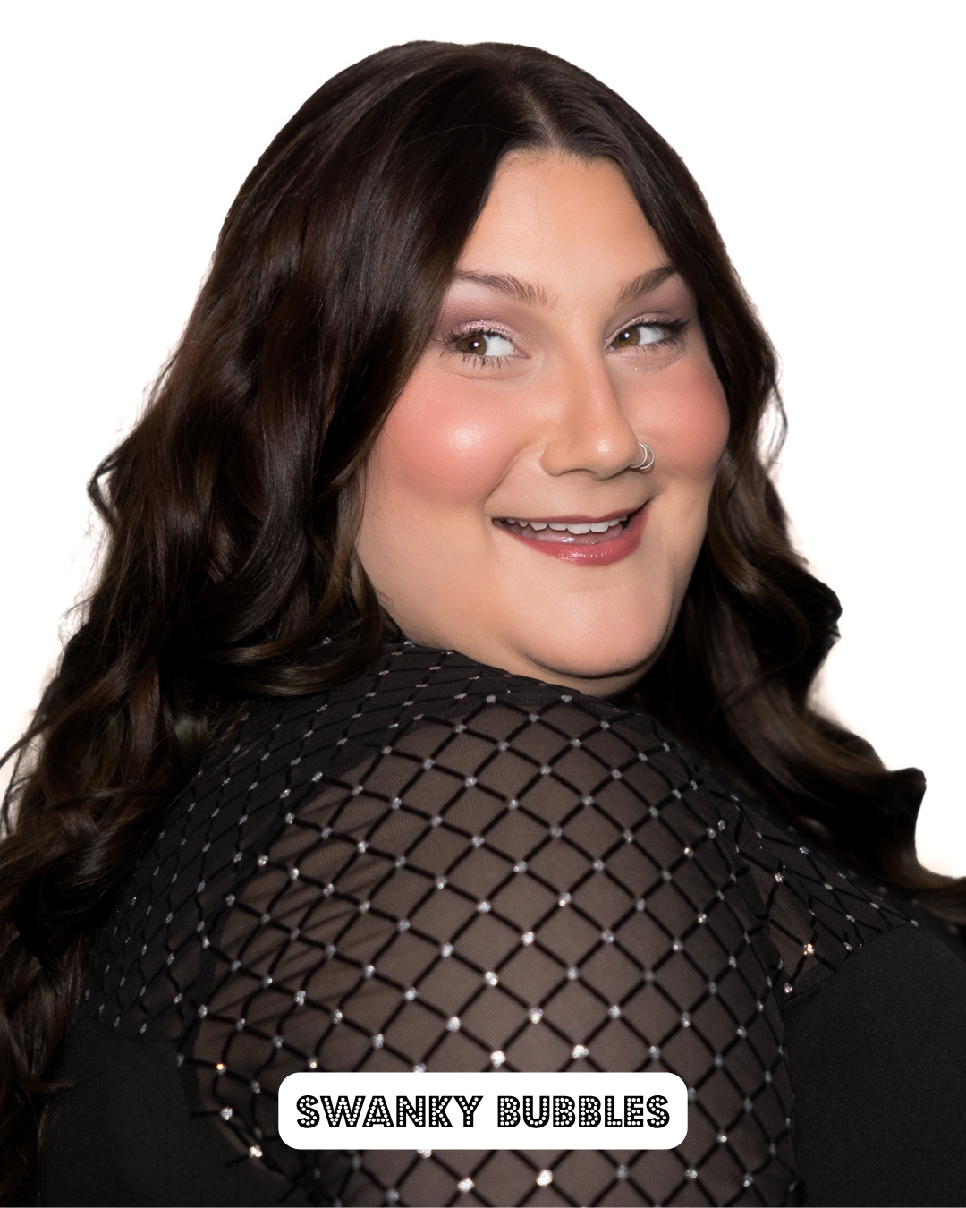 A woman with long, dark, wavy hair wearing a black, patterned top with small shiny accents, smiling and looking over her shoulder.