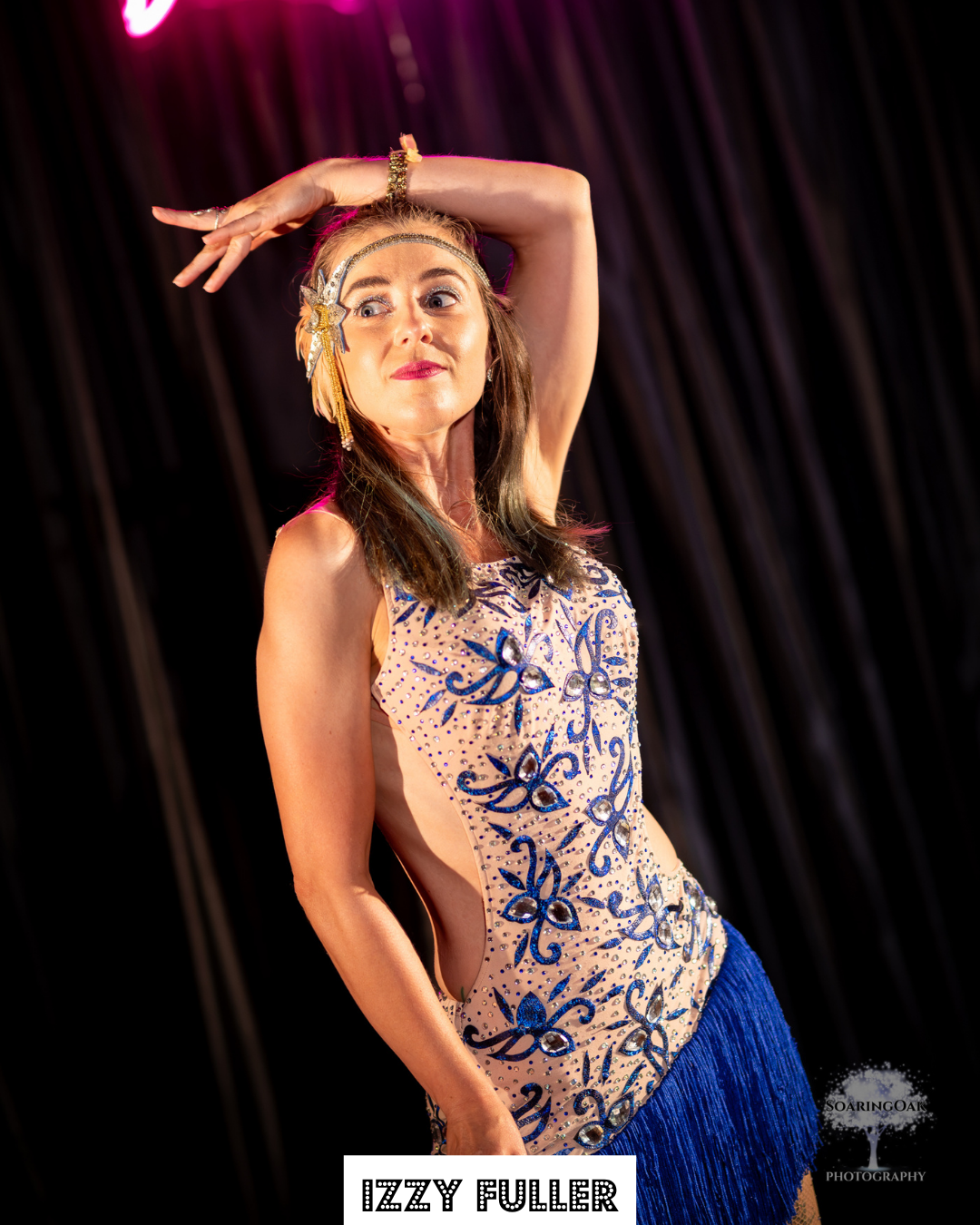 A woman with long brown hair and fair skin, dressed in a sparkly, floral-patterned dress with blue accents, performing a dance move on stage with her right arm raised and above her head. She is wearing a headband and jewelry, and the background is dark with stage lighting.