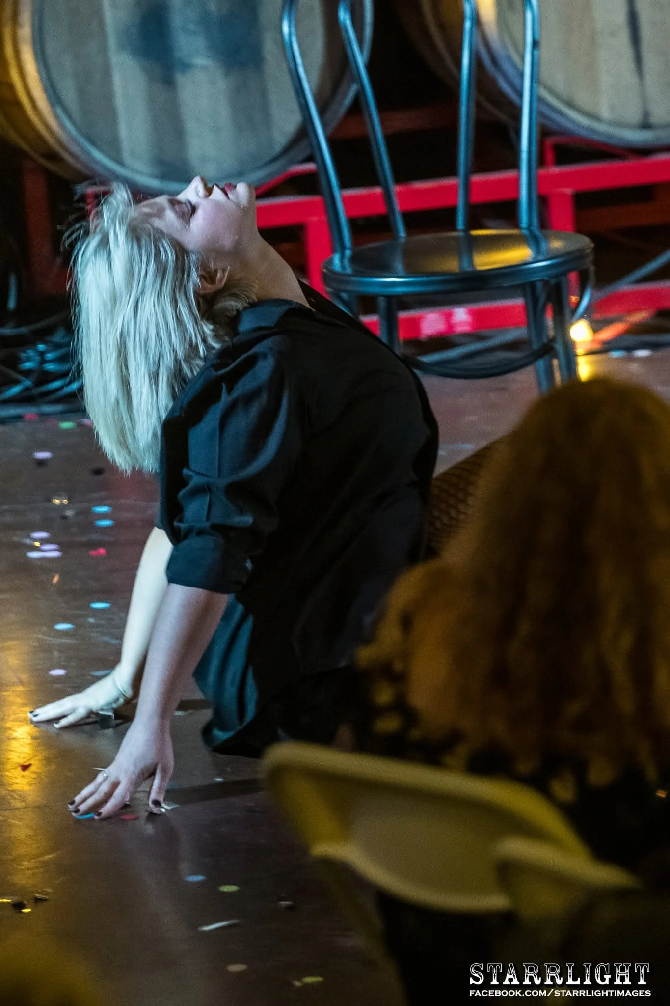 A woman with long platinum hair in a black outfit is performing a dance move on the floor with her hands and feet on the ground, head tilted back, in a dimly lit room with chairs stacked against the wall and confetti scattered on the floor.