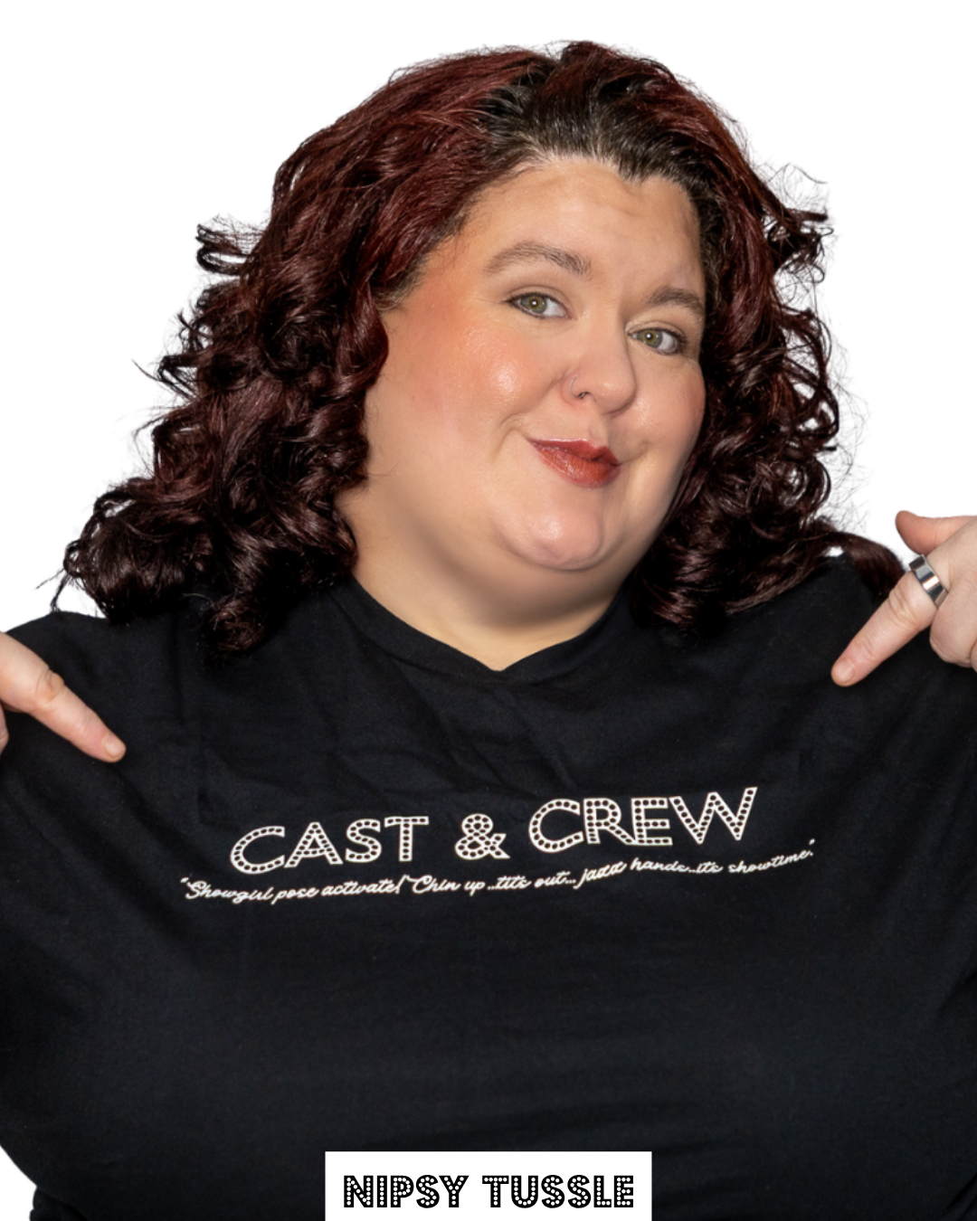 A woman with curly red hair and green eyes holding a black T-shirt that says 'CAST & CREW' and a quote below. She is smiling and pointing to the T-shirt with both hands. The background is white.