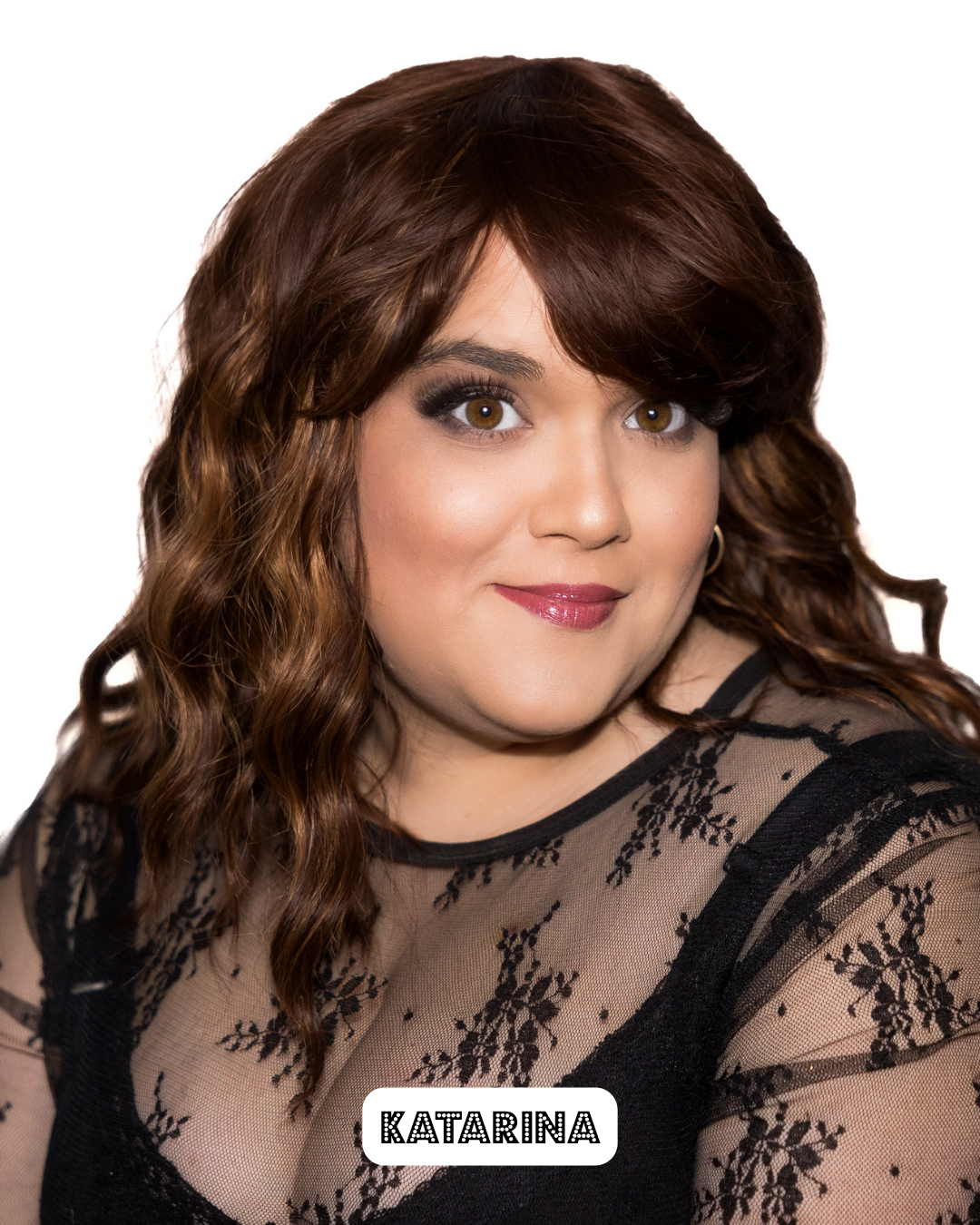 A woman with brown, wavy hair and brown eyes, wearing makeup with dark eyeshadow and pink lipstick, smiling at the camera. She is dressed in a black lace top with floral embroidery.
