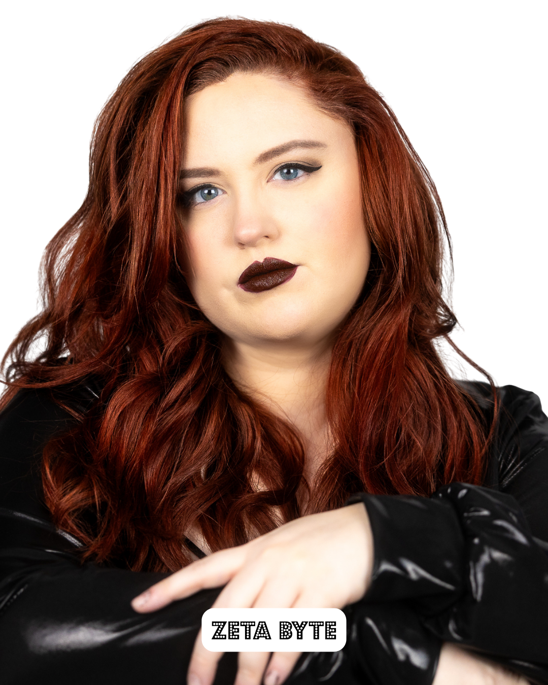 A woman with long, wavy red hair and blue eyes, wearing black makeup and a black leather jacket, looking at the camera with her arms crossed and a neutral expression, with the text 'ZETA BYTE' at the bottom.