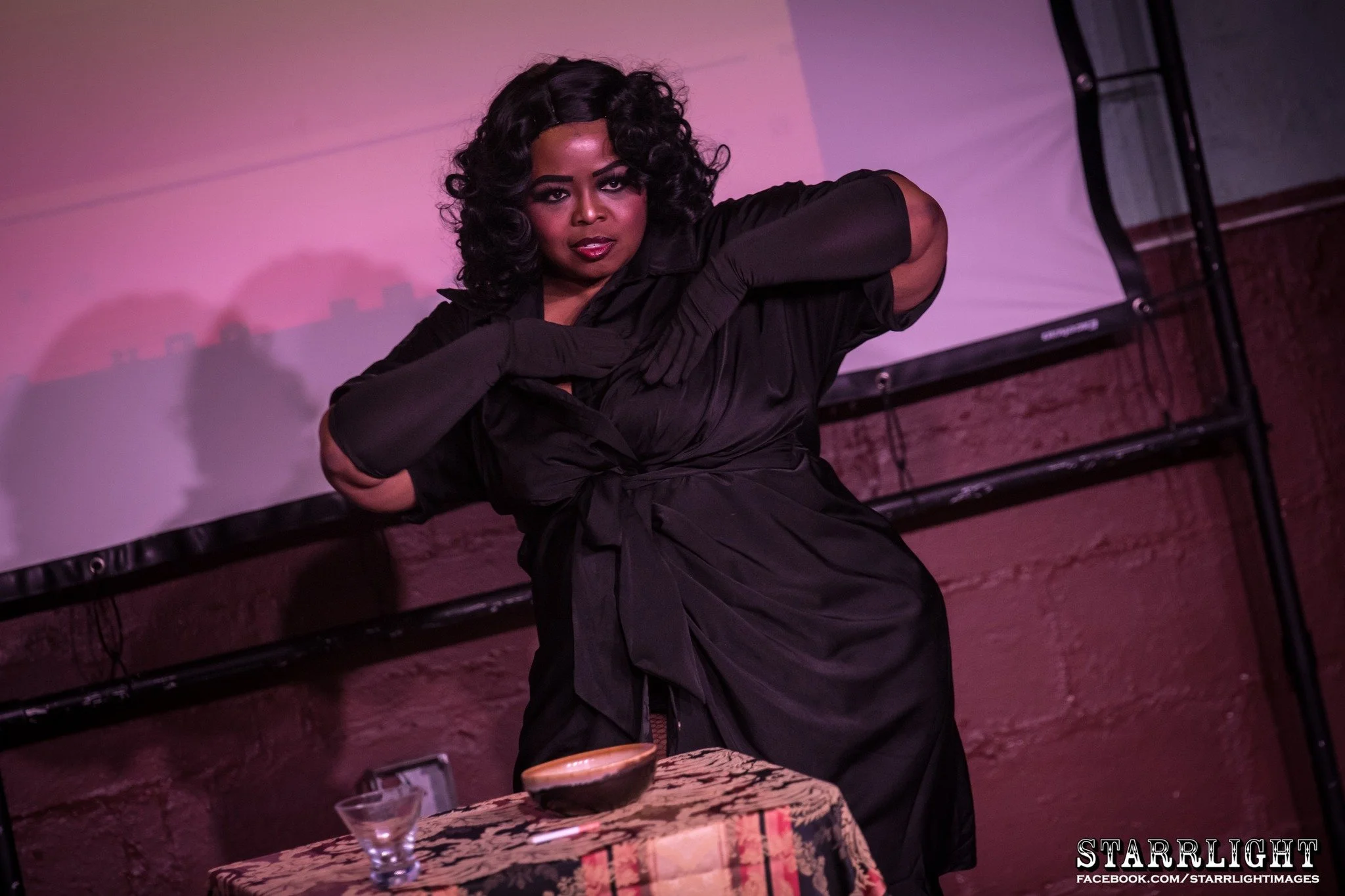 A person with dark, curly hair posing in front of a pinkish background, dressed in black, with a table and drinks in the foreground.