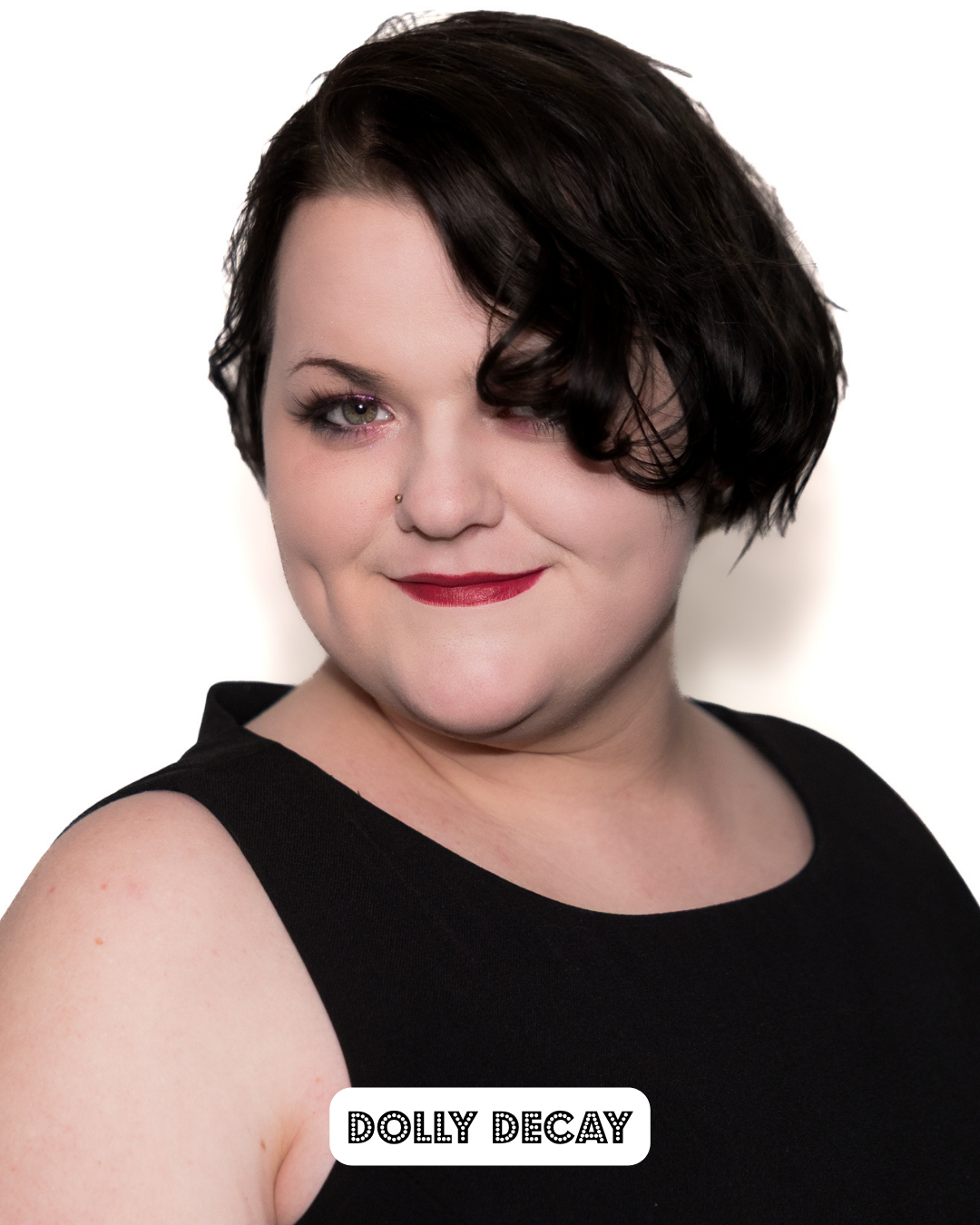 Portrait of a woman with short dark hair, wearing a black sleeveless top, red lipstick, and a nose piercing, smiling slightly at the camera with a white background.