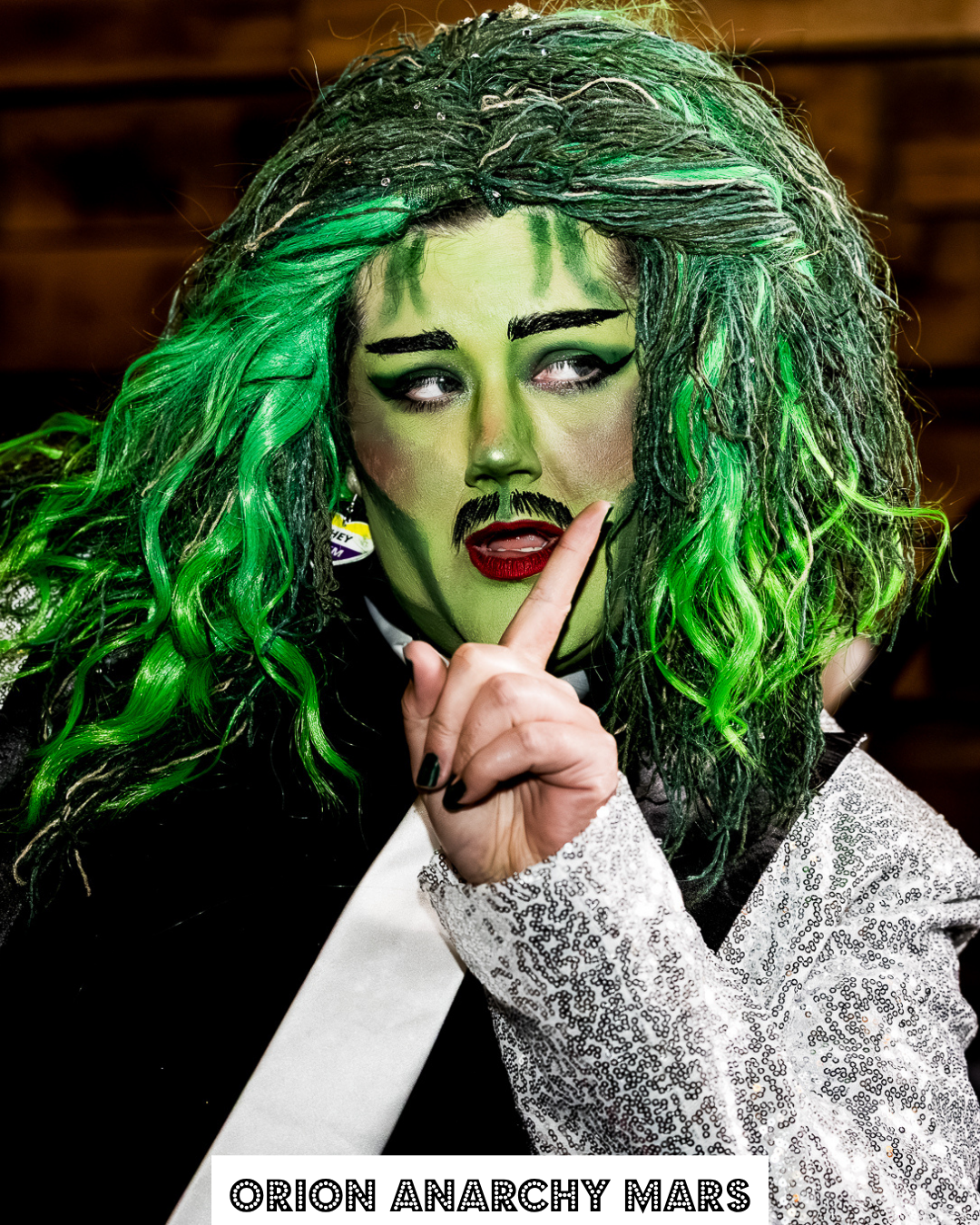 Person dressed as a fictional character with green makeup, green and black curly hair, dark eye makeup, red lipstick, and a drawn-on mustache. They are holding a finger to their lips in a shushing gesture and wearing a white and black sequined outfit. The text at the bottom reads "ORION ANARCHY MARS."