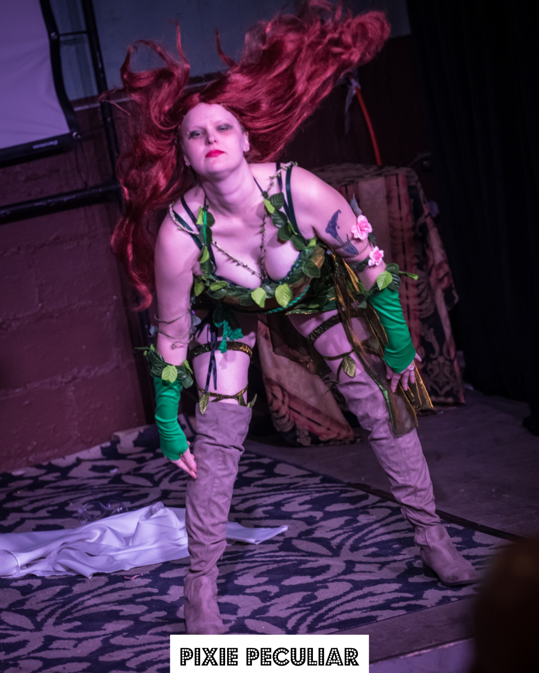A woman with long red hair, dressed in an elaborate costume with green leaves, pink flowers, and gray boots, performing on stage under pink lighting.