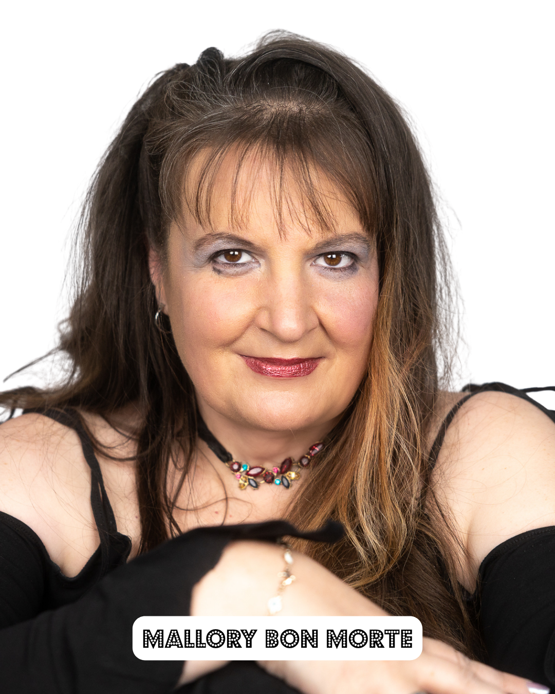 Portrait of Mallory Bon Morte with long brown hair, wearing black off-shoulder top, colorful necklace, and makeup, smiling softly against a white background.