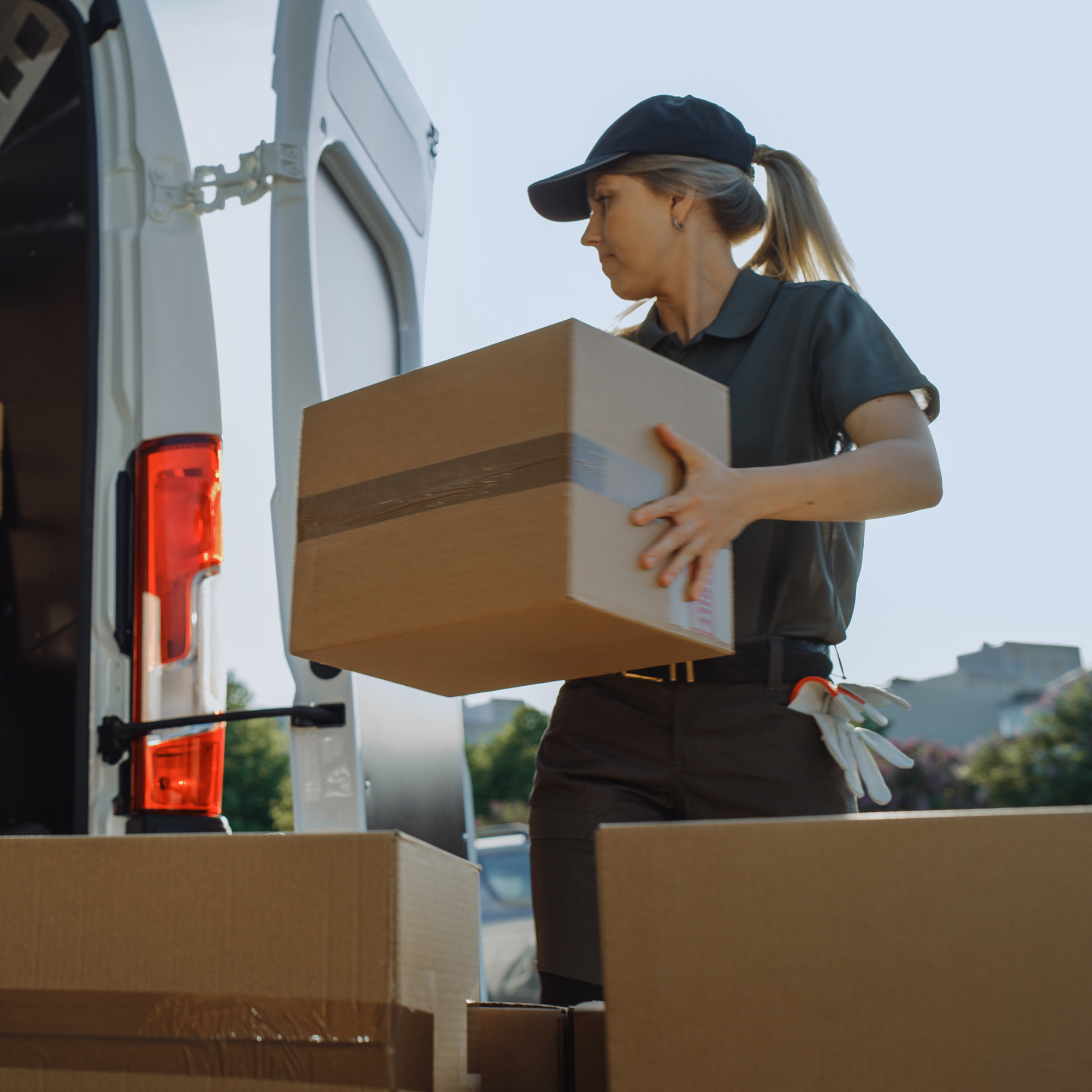A woman wearing a black cap and dark clothing is loading a cardboard box into the back of a white van outdoors during daytime.