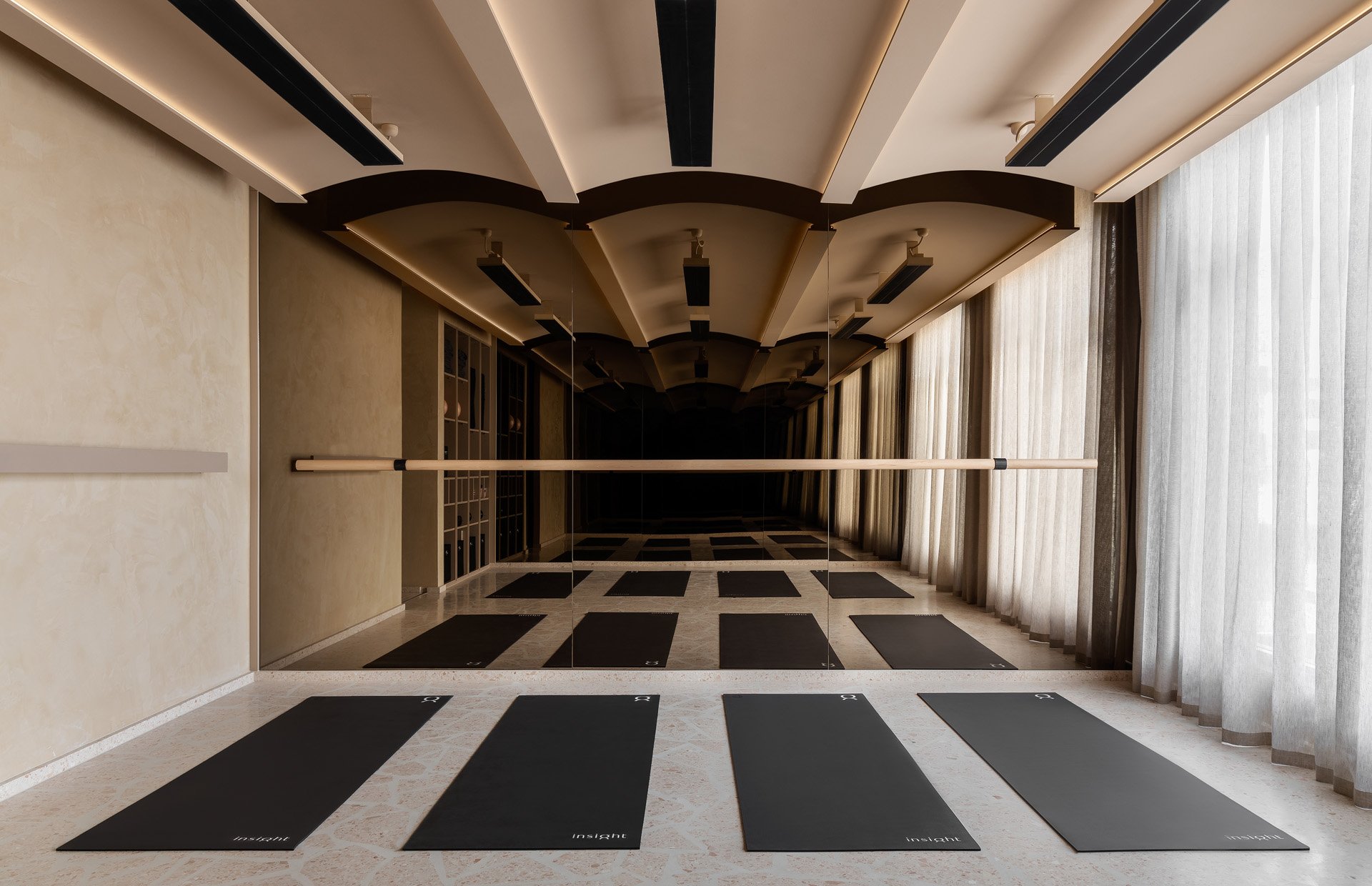 A yoga studio with black mats arranged in a row on tiled floor. The room has beige walls, a large mirror covering the entire wall, and floor-to-ceiling curtains on the right side. The ceiling has curved architectural features with lighting and black rectangular hanging lights.