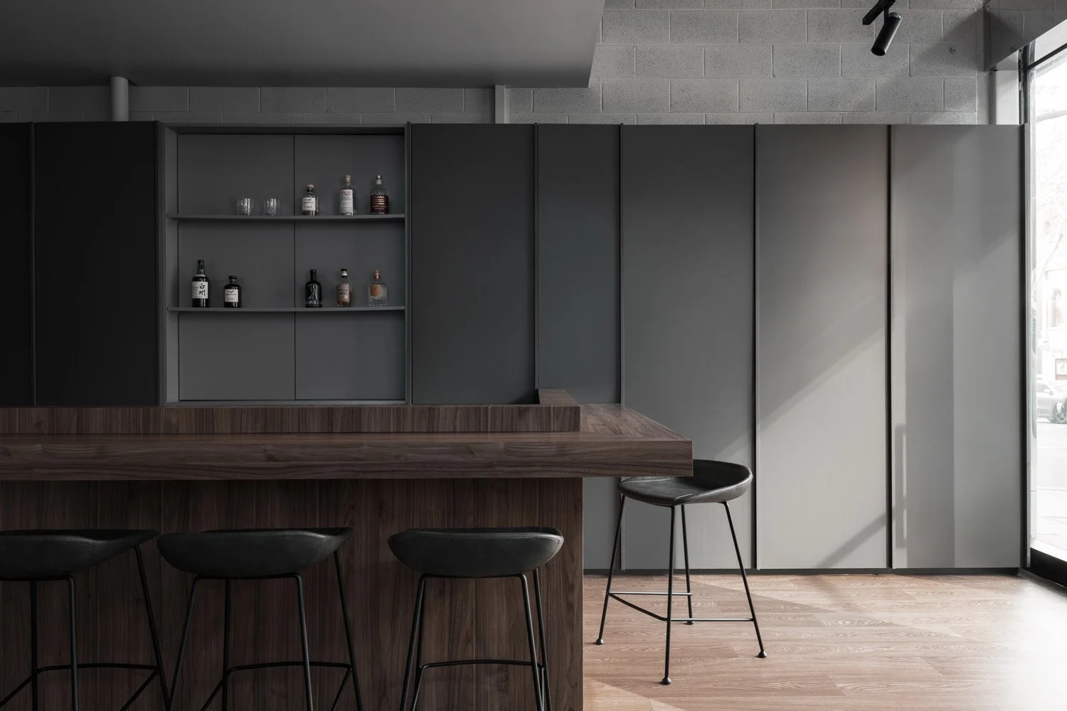 Modern interior with a wooden bar counter, black bar stools, and dark gray cabinets, with bottles and glasses on open shelves, natural light coming from a door on the right.