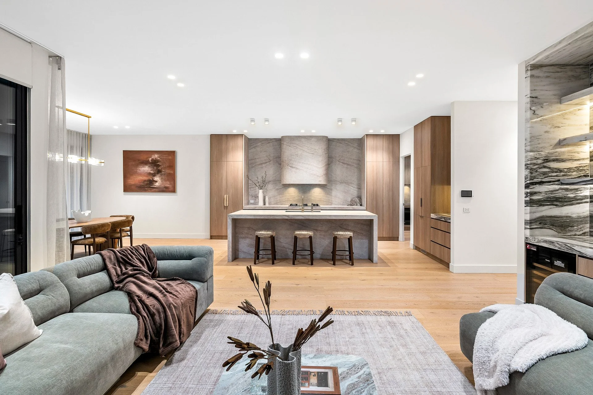 Open-concept living room and kitchen with modern decor, light wood flooring, grey sofa, armchair with throw blanket, wooden dining table and chairs, marble accent wall, and minimalist design.