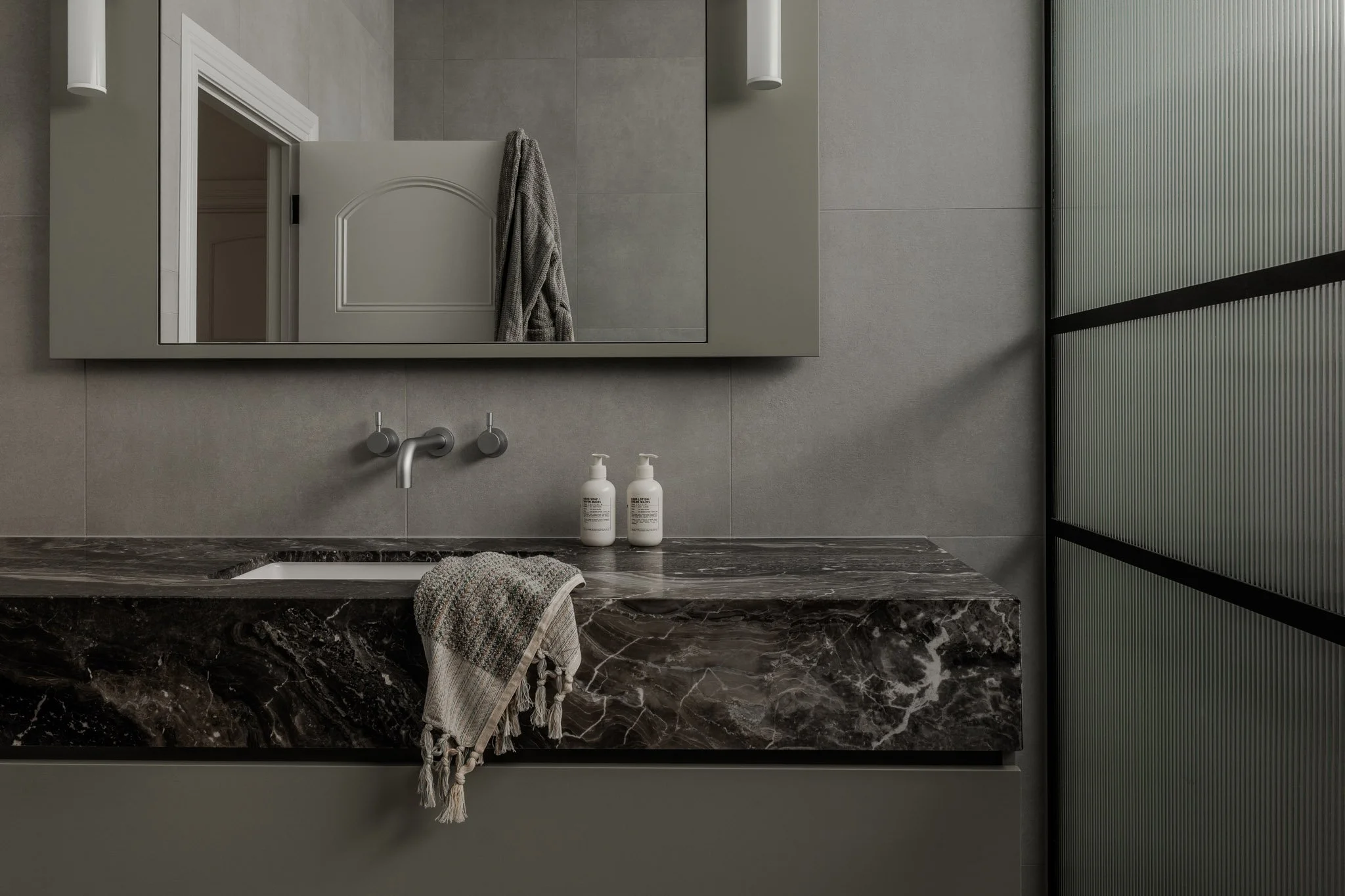 Modern bathroom with a large marble countertop, a wall-mounted round faucet, a mirror, two bottles of soap, a towel, and a textured glass panel.