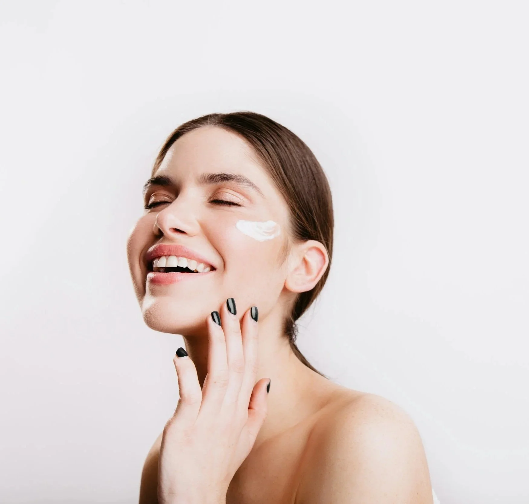 A woman smiling with her eyes closed has a dollop of white facial cream on her cheek, and her fingers with black nail polish gently touching her face.