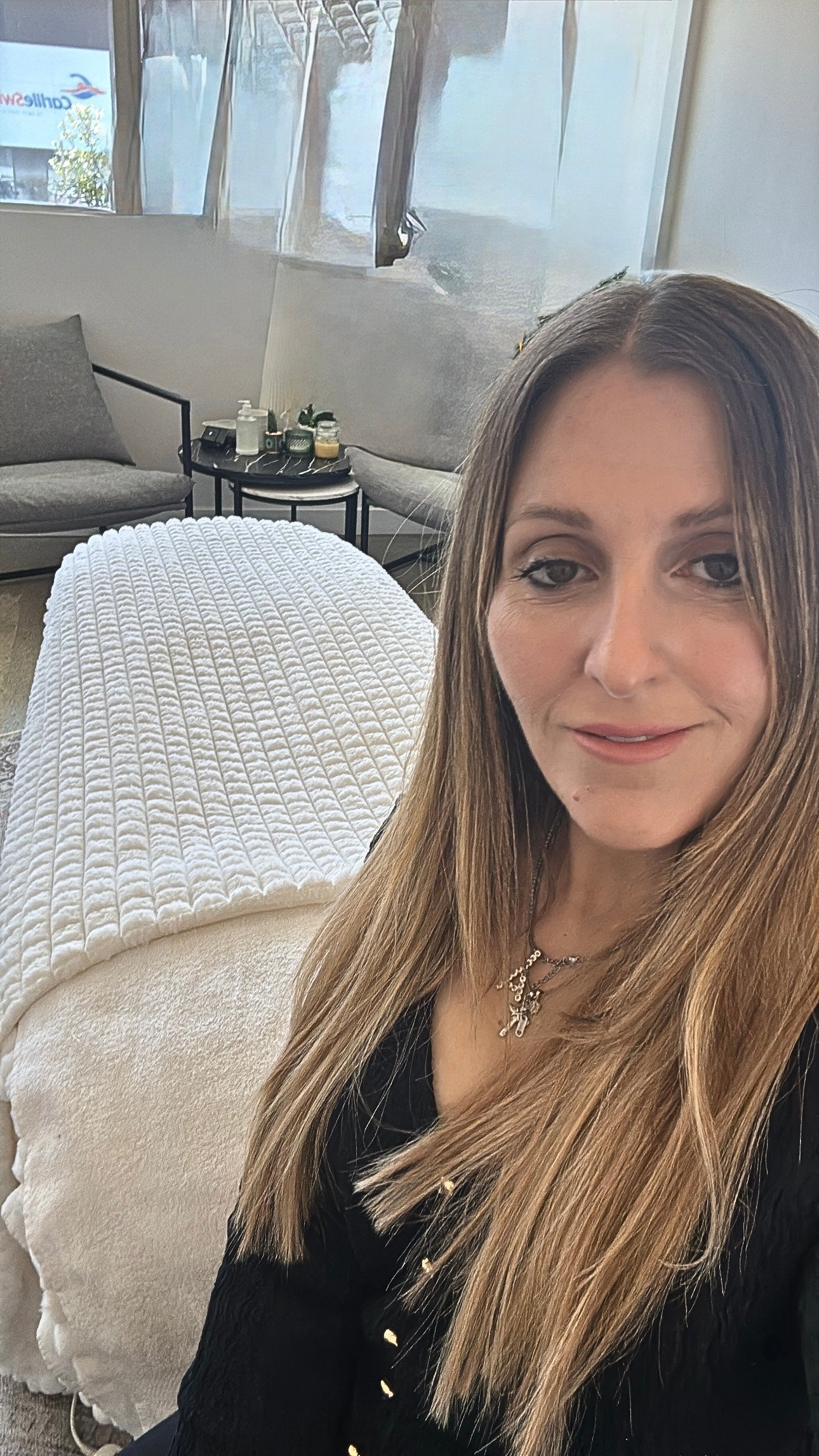 A woman with long light brown hair, wearing a black outfit and a cross necklace, sitting on a beige textured sofa in a room with large windows and modern decor.