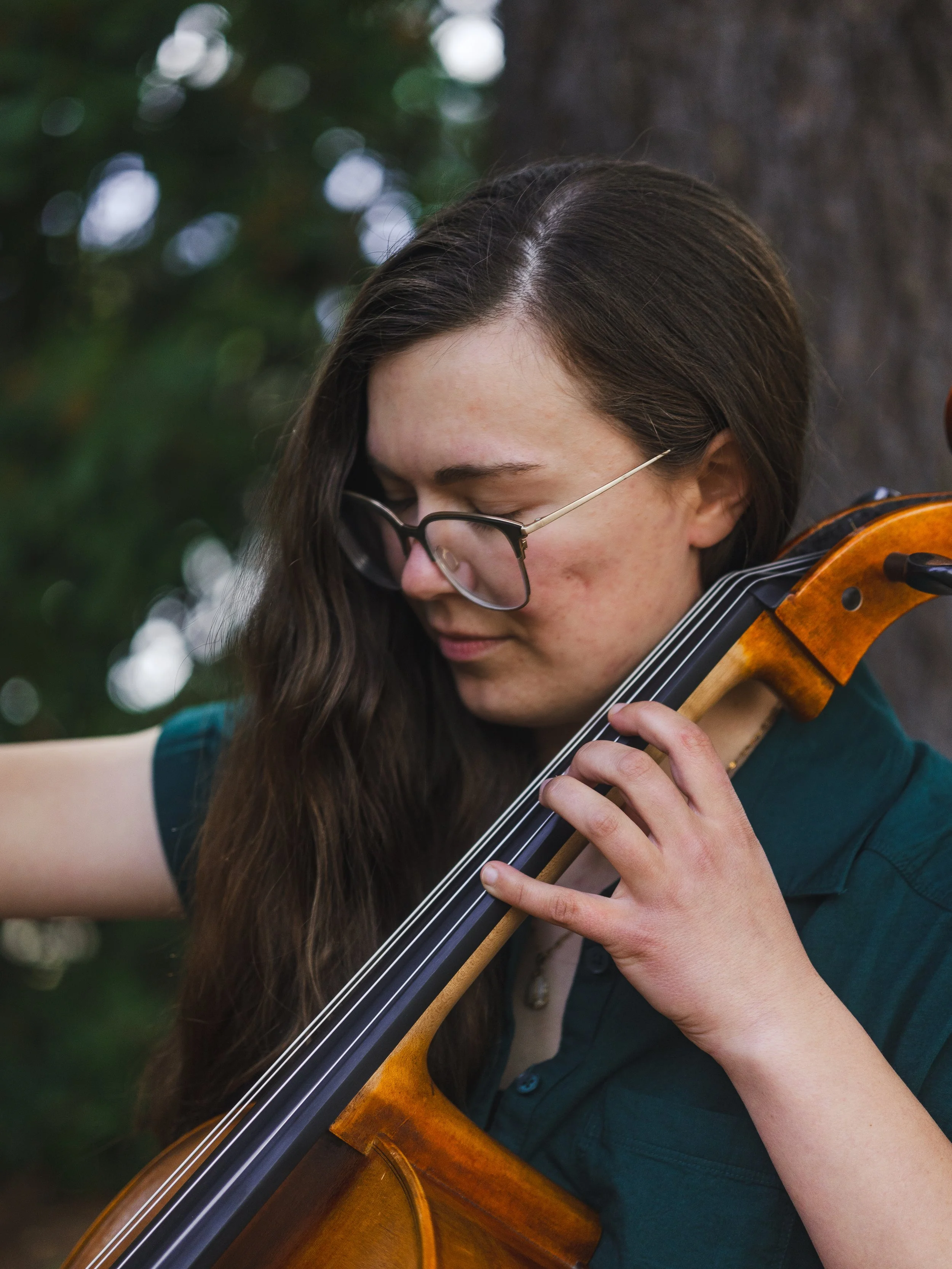 A young woman with long brown hair, glasses, and a teal shirt playing a violin outdoors in front of a blurred green and white bokeh background.