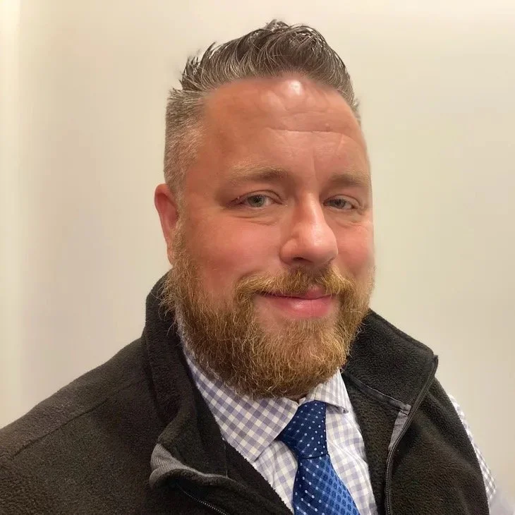 A man with a beard and short hair, wearing a checkered shirt, a blue tie, and a black jacket, smiling at the camera against a plain background.