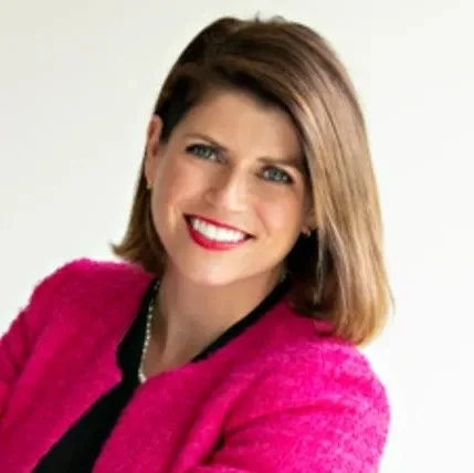 A woman with shoulder-length brown hair smiling, wearing a pink blazer and a black top, against a light background.